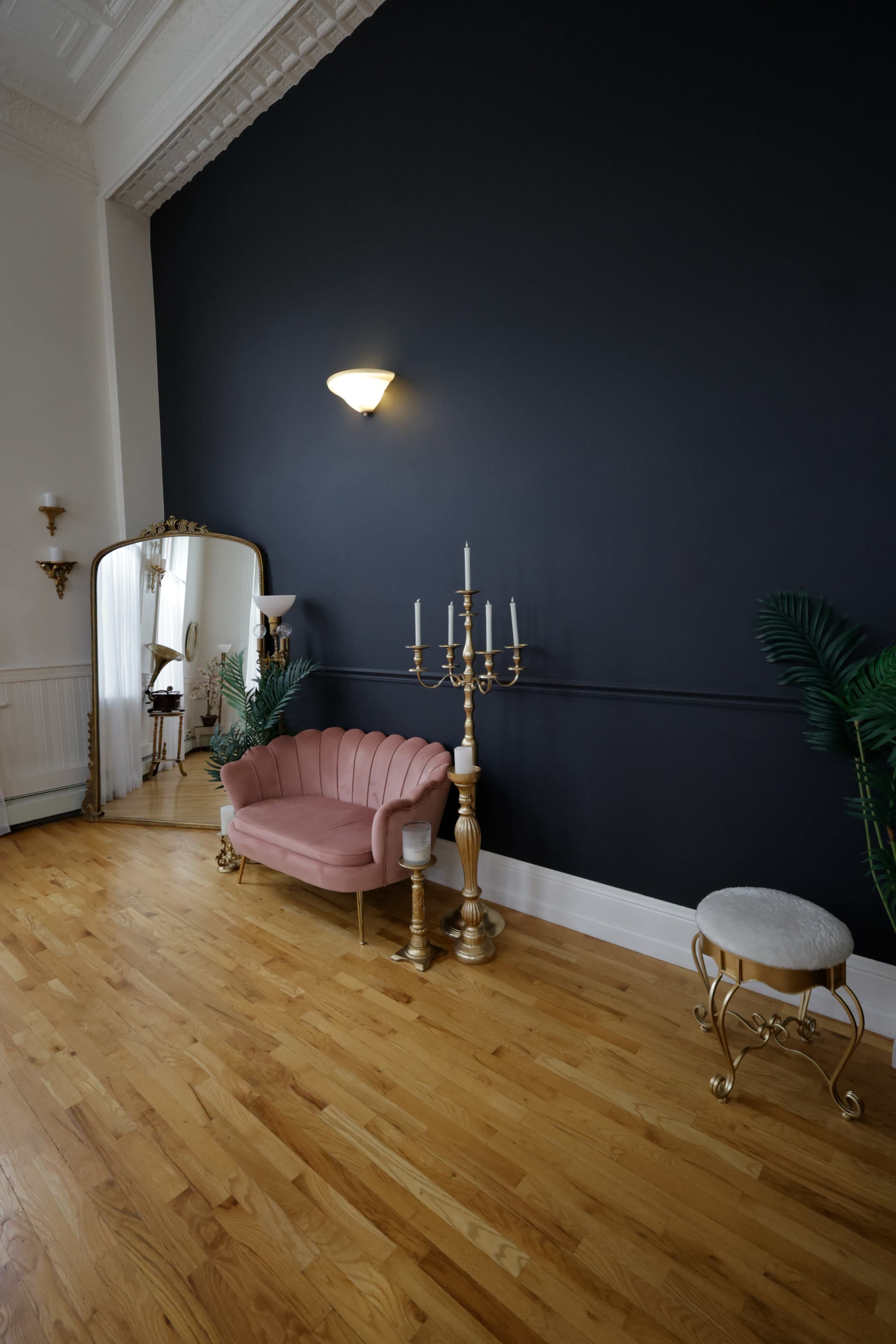 The scene features a pink velvet loveseat, a gold candelabrum, a round stool, and a tall mirror against a dark blue wall in a room with wooden flooring.