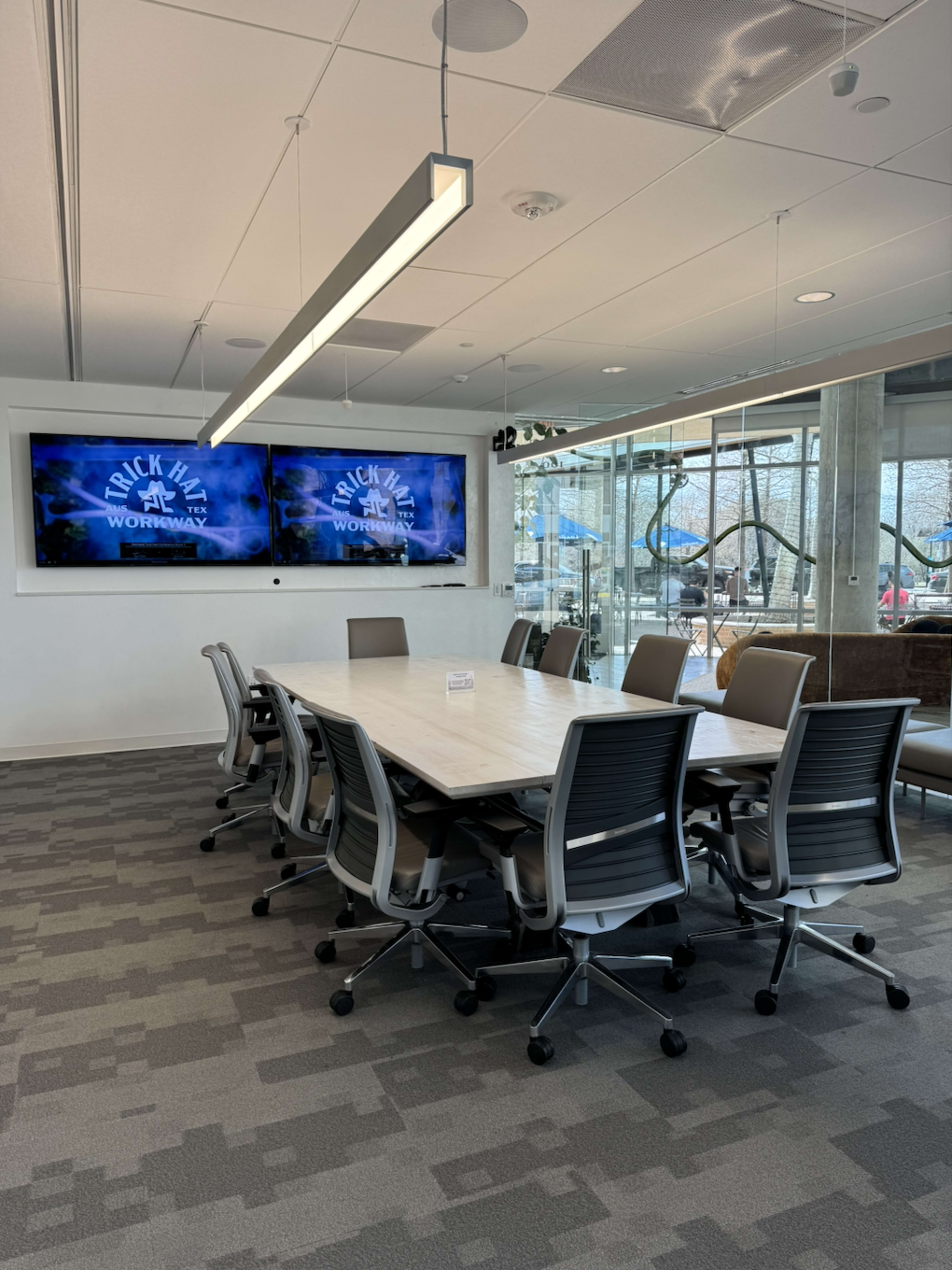 The image shows a modern conference room featuring a large table surrounded by rolling chairs, two wall-mounted screens displaying a graphic, and large windows allowing natural light.