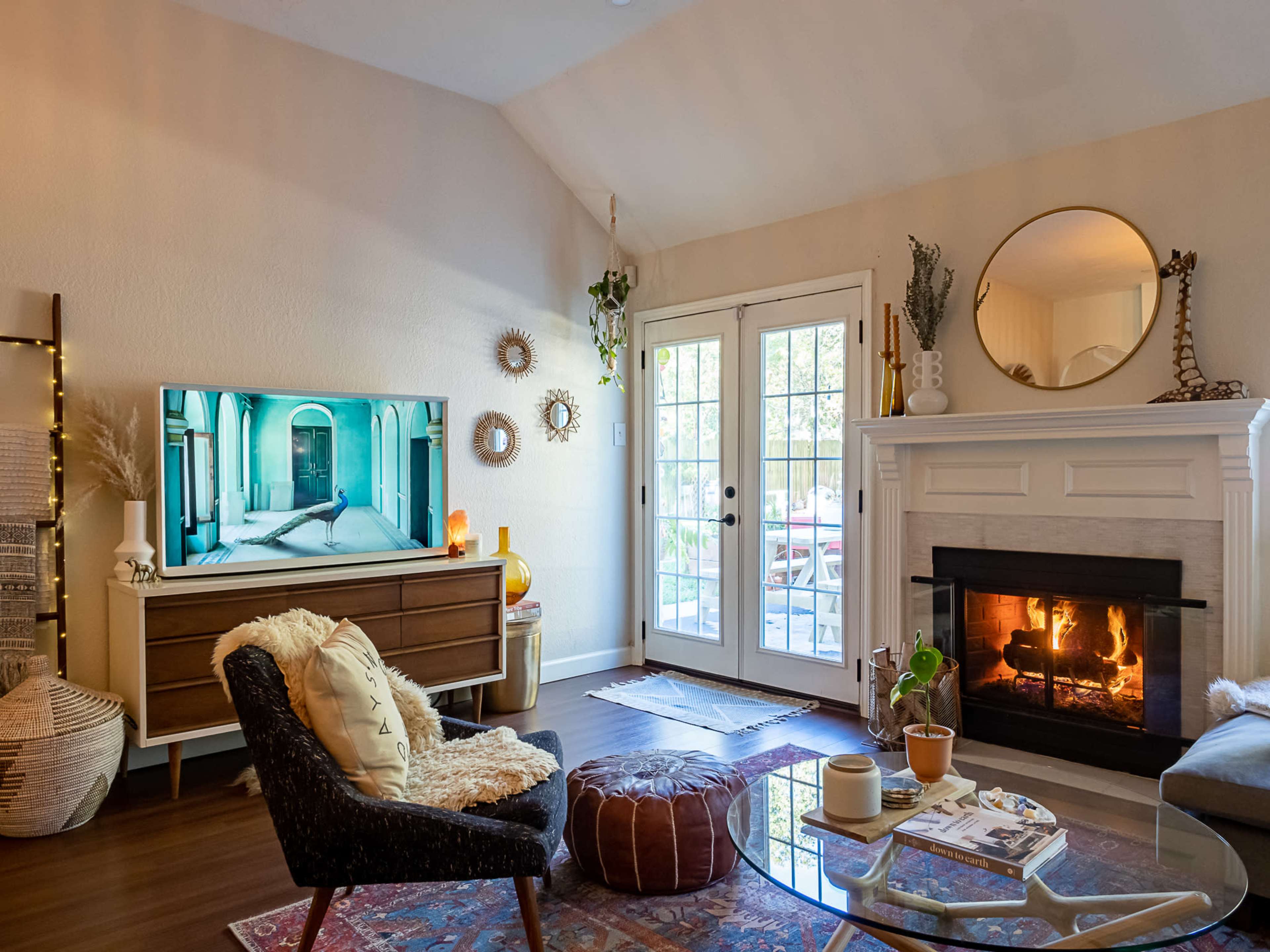 A modern living room features a TV mounted on a wall, a cozy seating arrangement with a chair and an ottoman, a fireplace with a burning fire, and glass doors leading to an outdoor patio.
