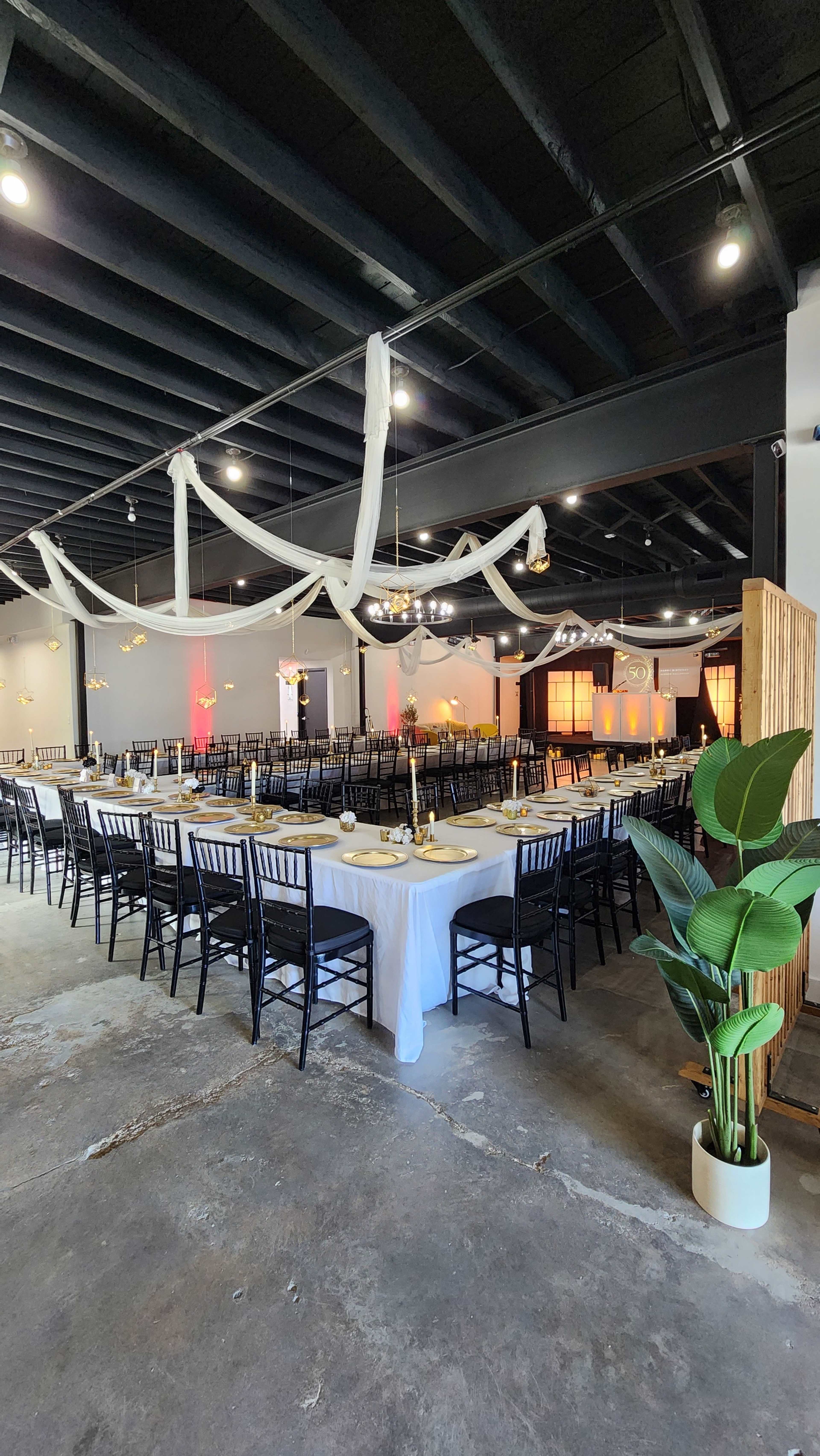 The image shows a spacious event venue with rows of black chairs set around a long white table, decorated with gold accents and draped fabric overhead.