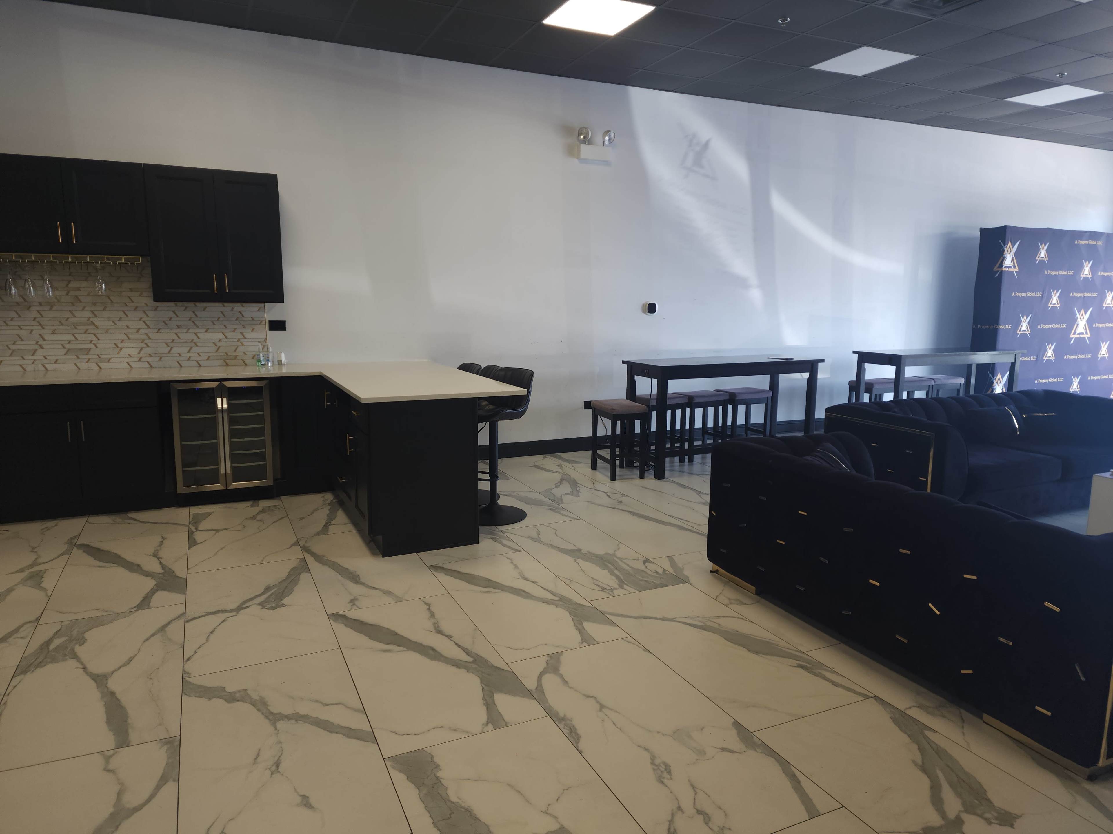 The image shows a modern indoor space with a kitchen area featuring black cabinetry and a bar, alongside multiple tables and chairs placed on a marble floor.