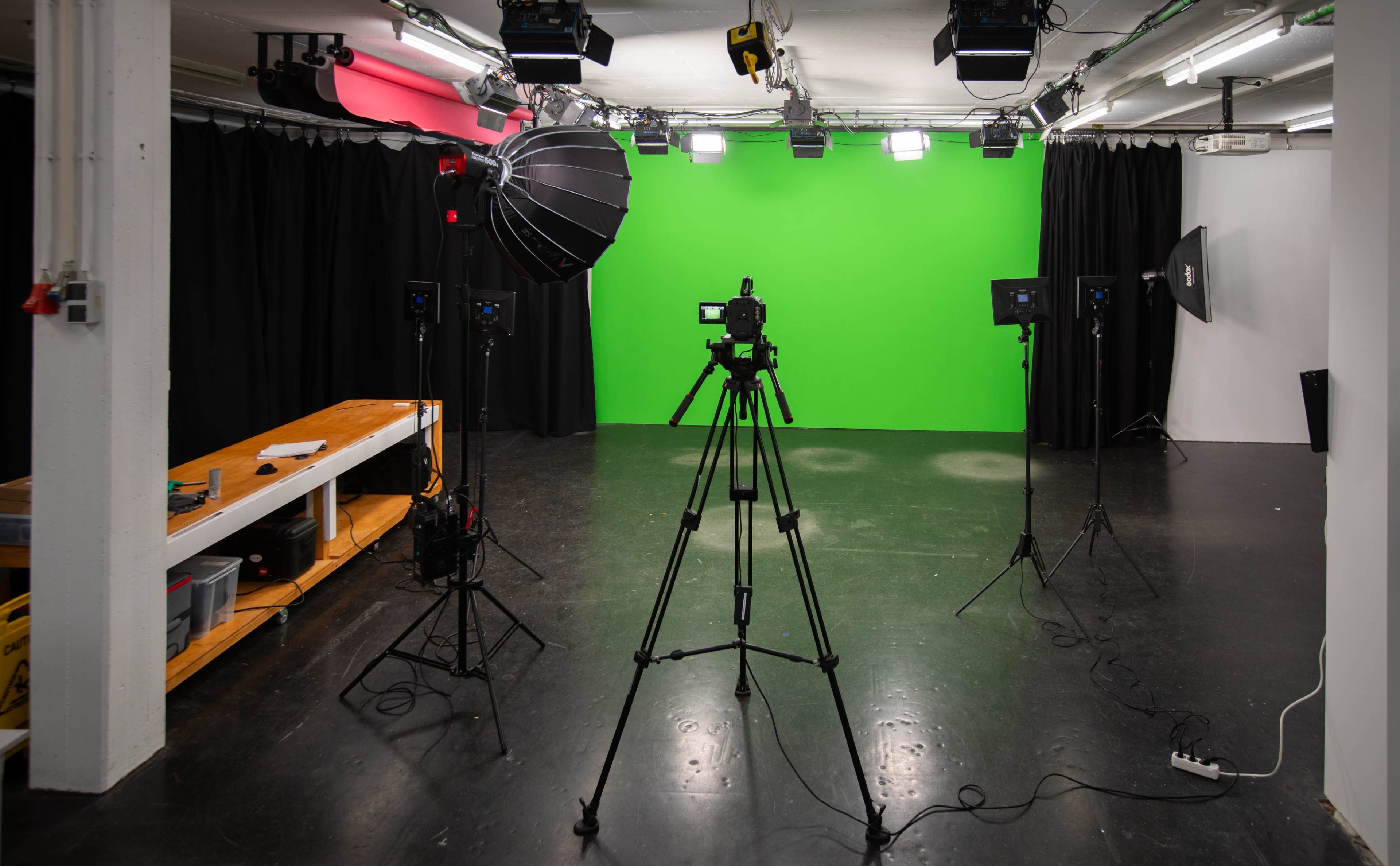 A photography studio features a green backdrop surrounded by multiple lights and tripods set up for video production.