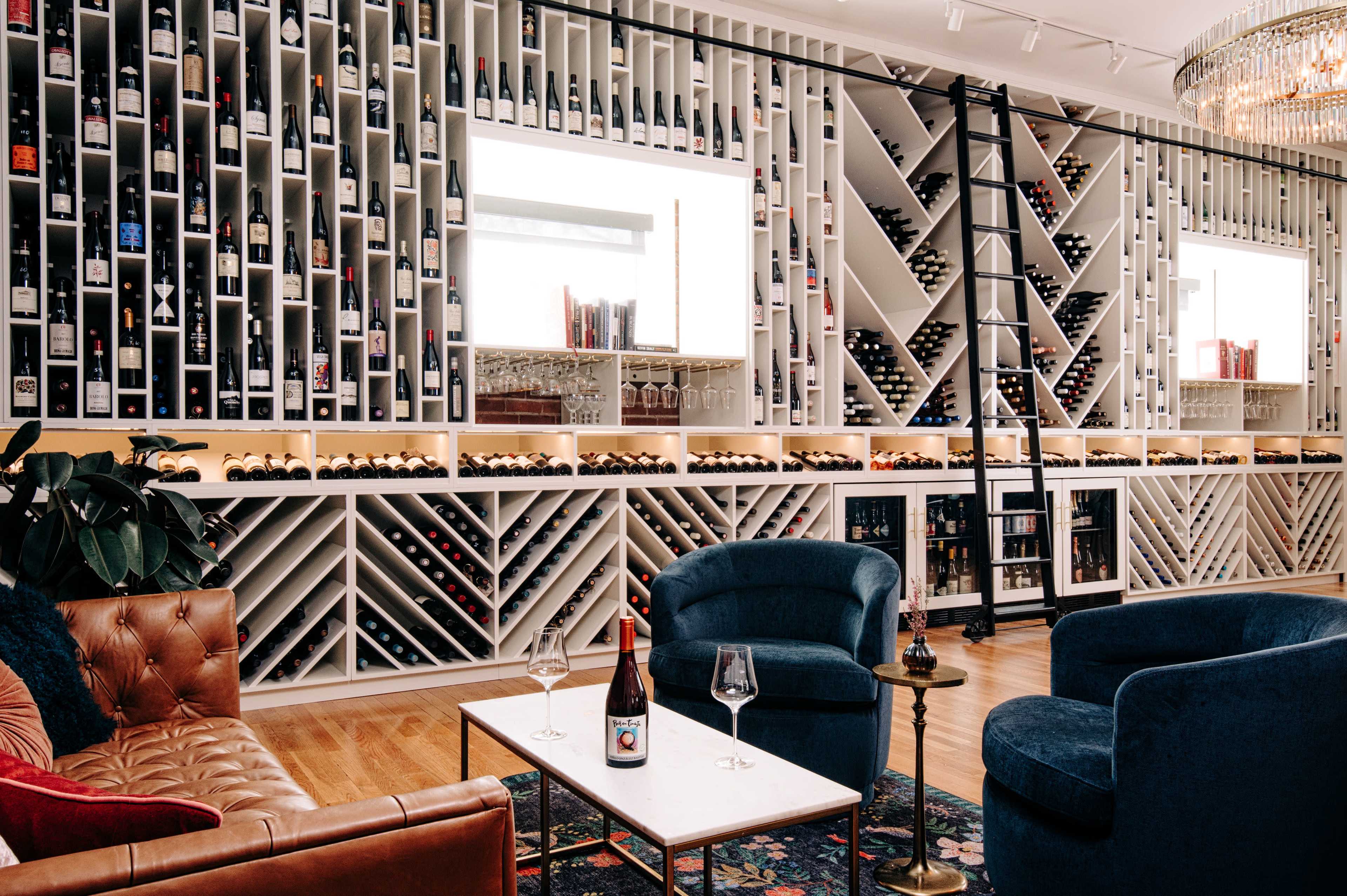 A modern wine display room with a wall of neatly arranged wine bottles and a cozy seating area featuring two chairs and a small table.