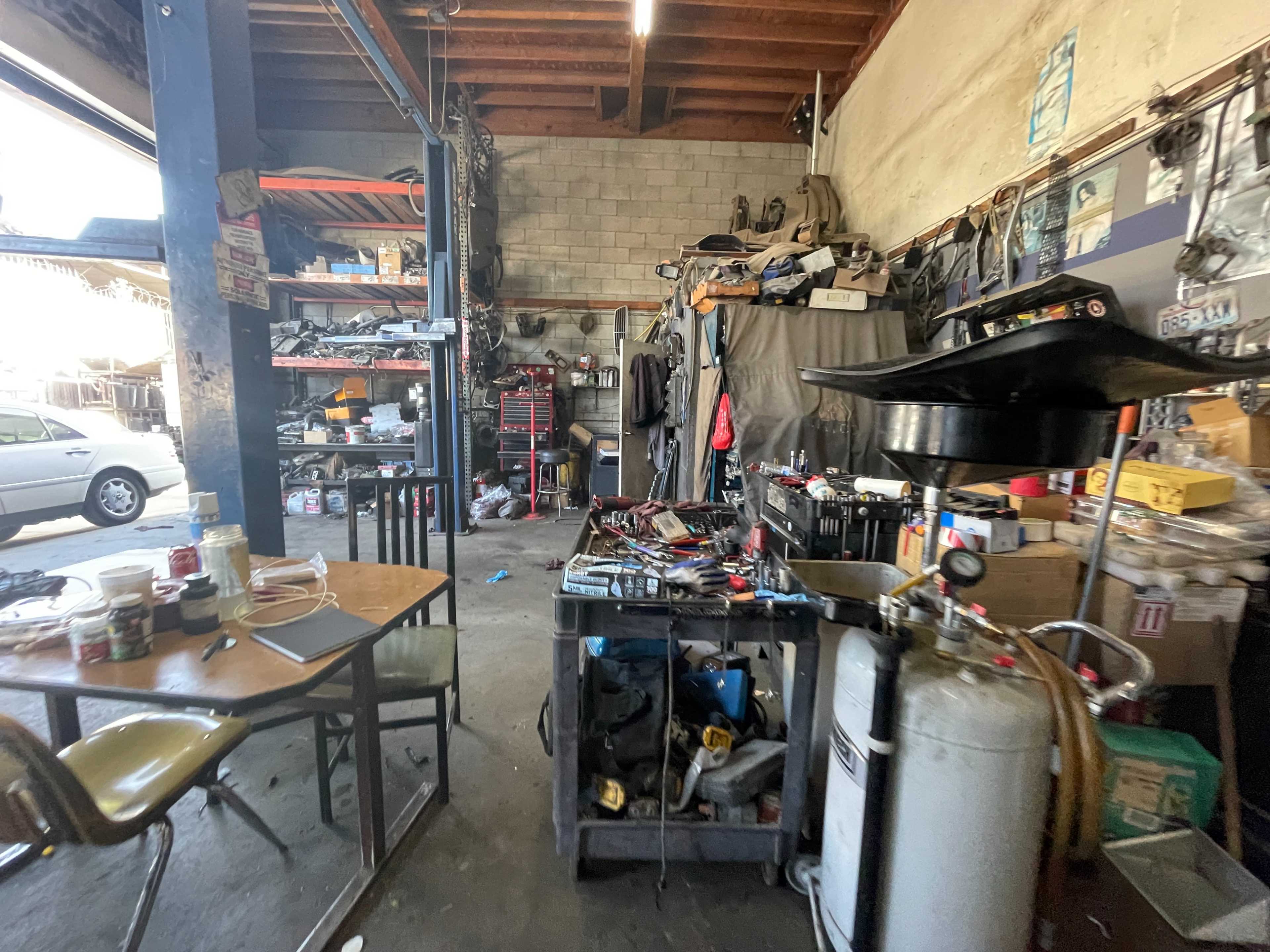 The image shows a cluttered garage or workshop filled with tools, parts, and equipment, with a table in the foreground and various items stacked on shelves.