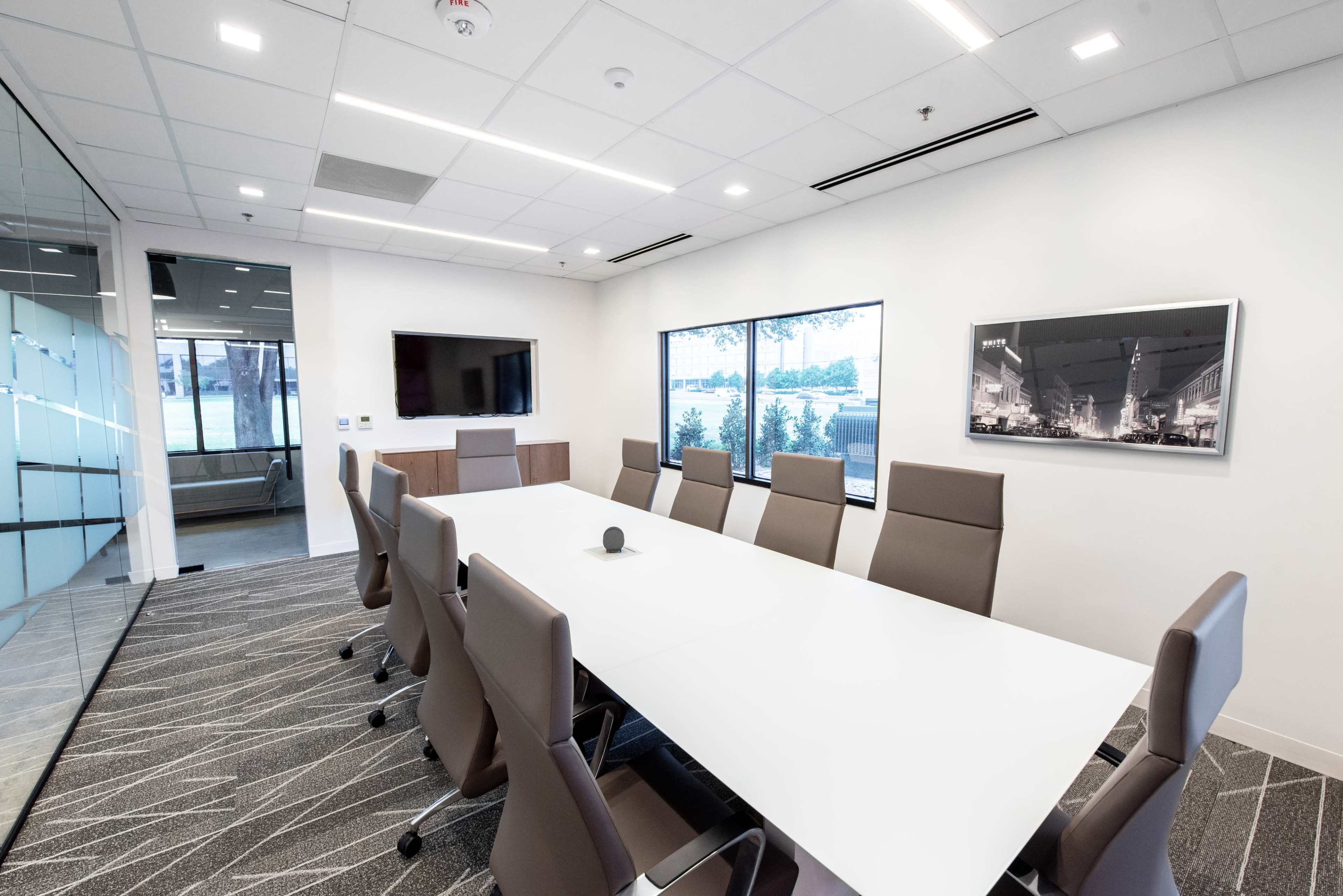 A modern conference room features a large white table surrounded by ergonomic chairs, with a television and large windows showcasing a view outside.