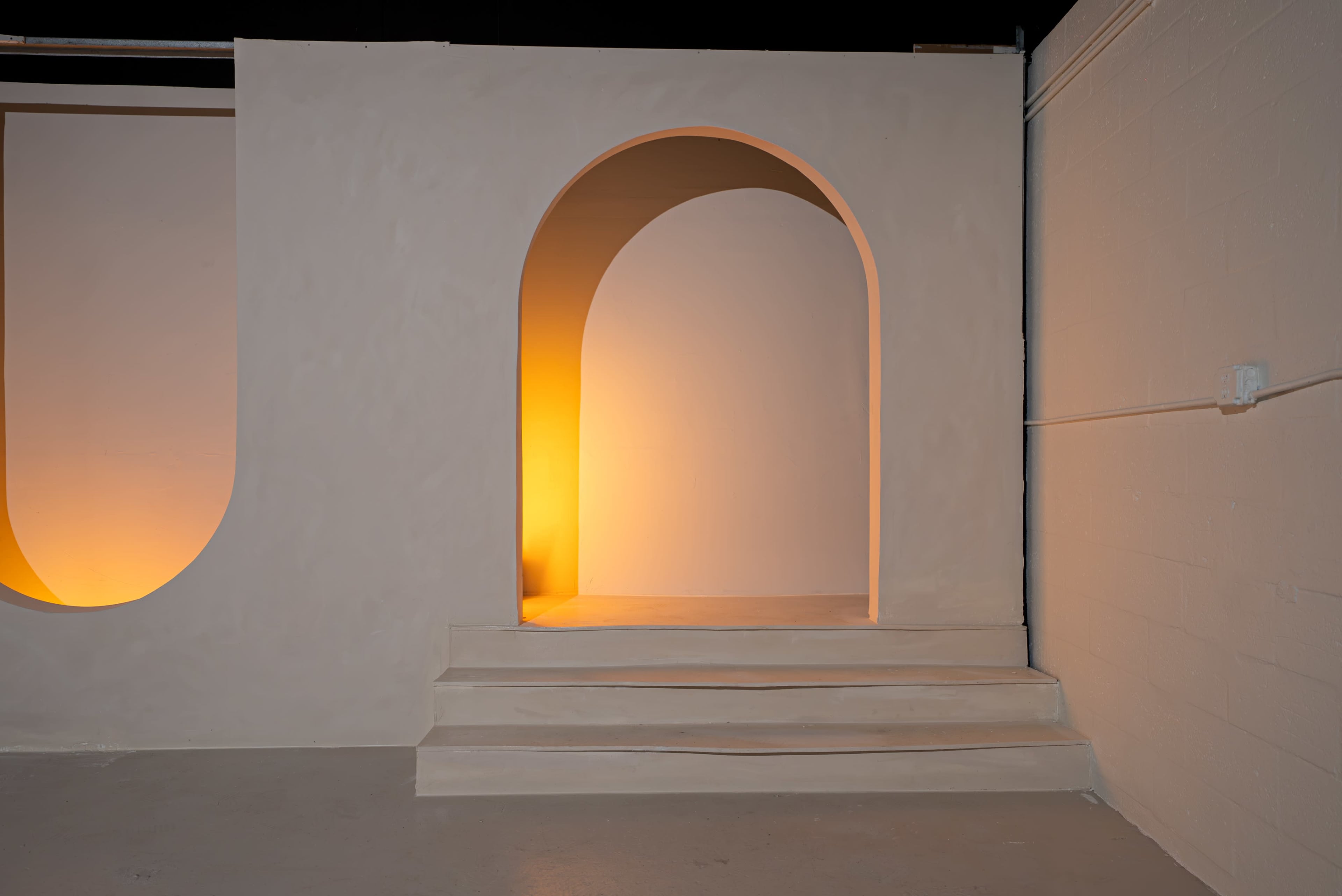 The image shows a minimalist architectural space featuring a wall with two arch-shaped cutouts, one illuminated with a warm light, and steps leading up to a raised platform.