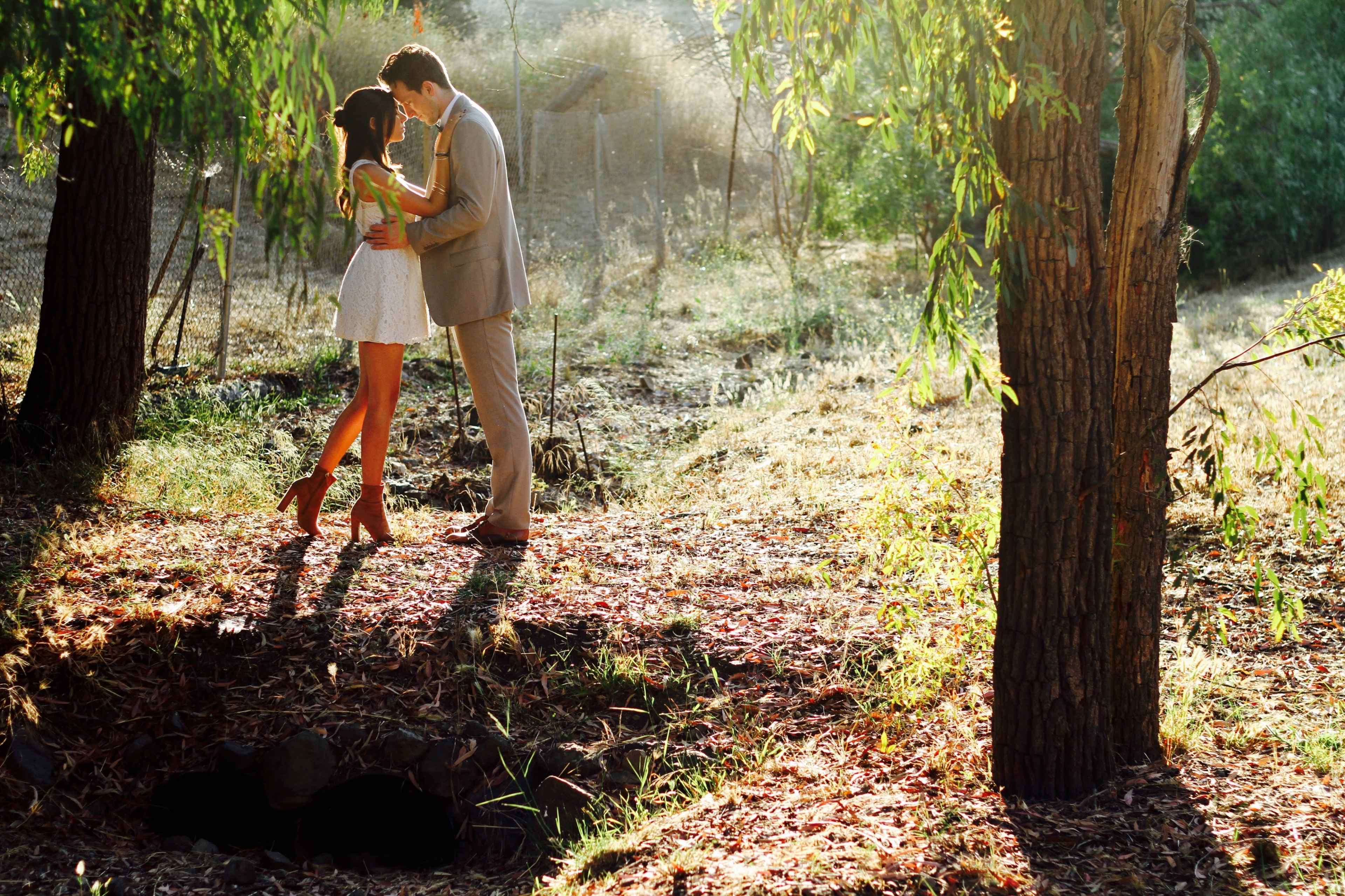 A couple stands closely together in a sunlit clearing surrounded by trees, sharing an intimate moment.