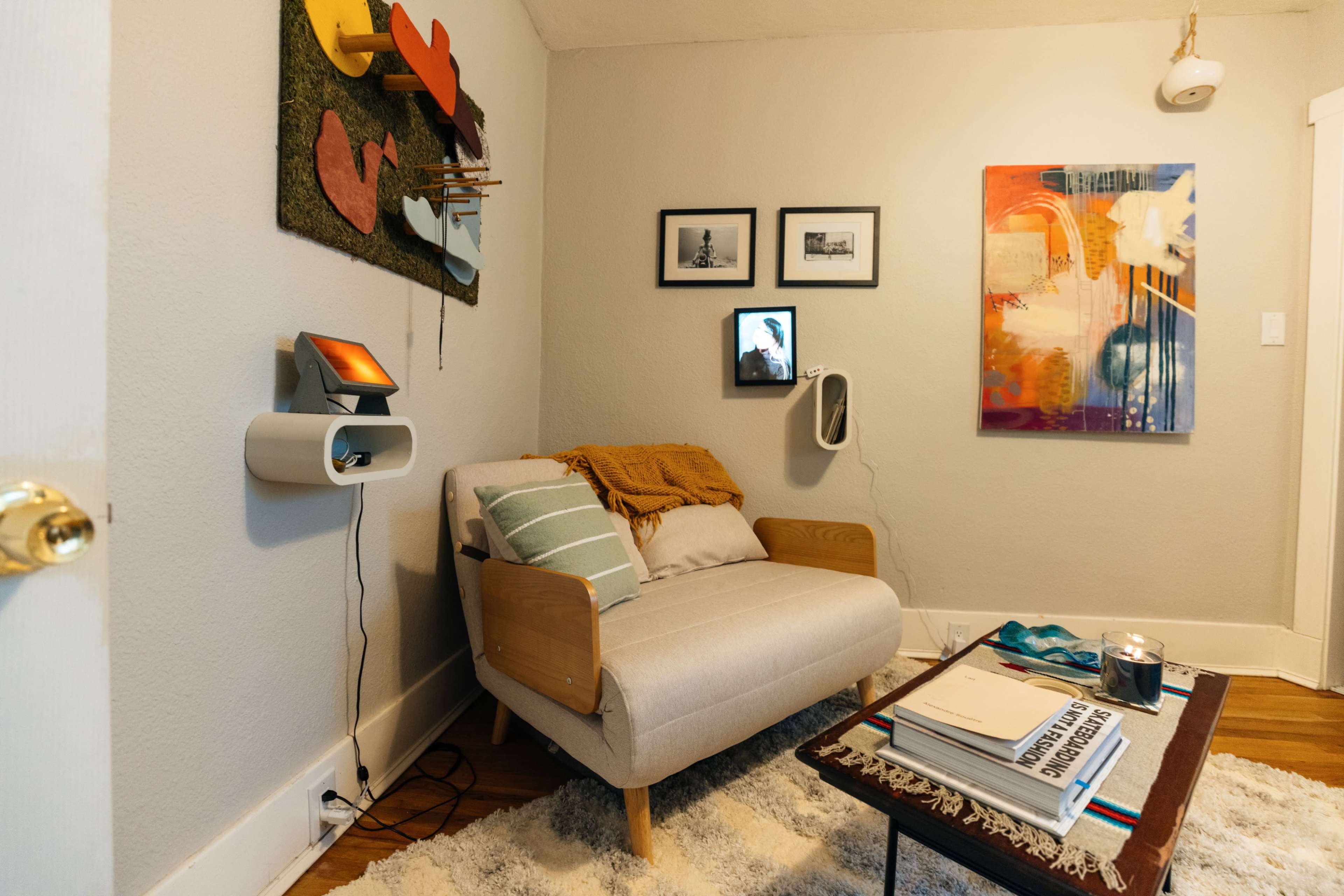 The image shows a cozy living room corner featuring a light-colored sofa, a coffee table with books and a candle, and colorful wall art alongside framed pictures.