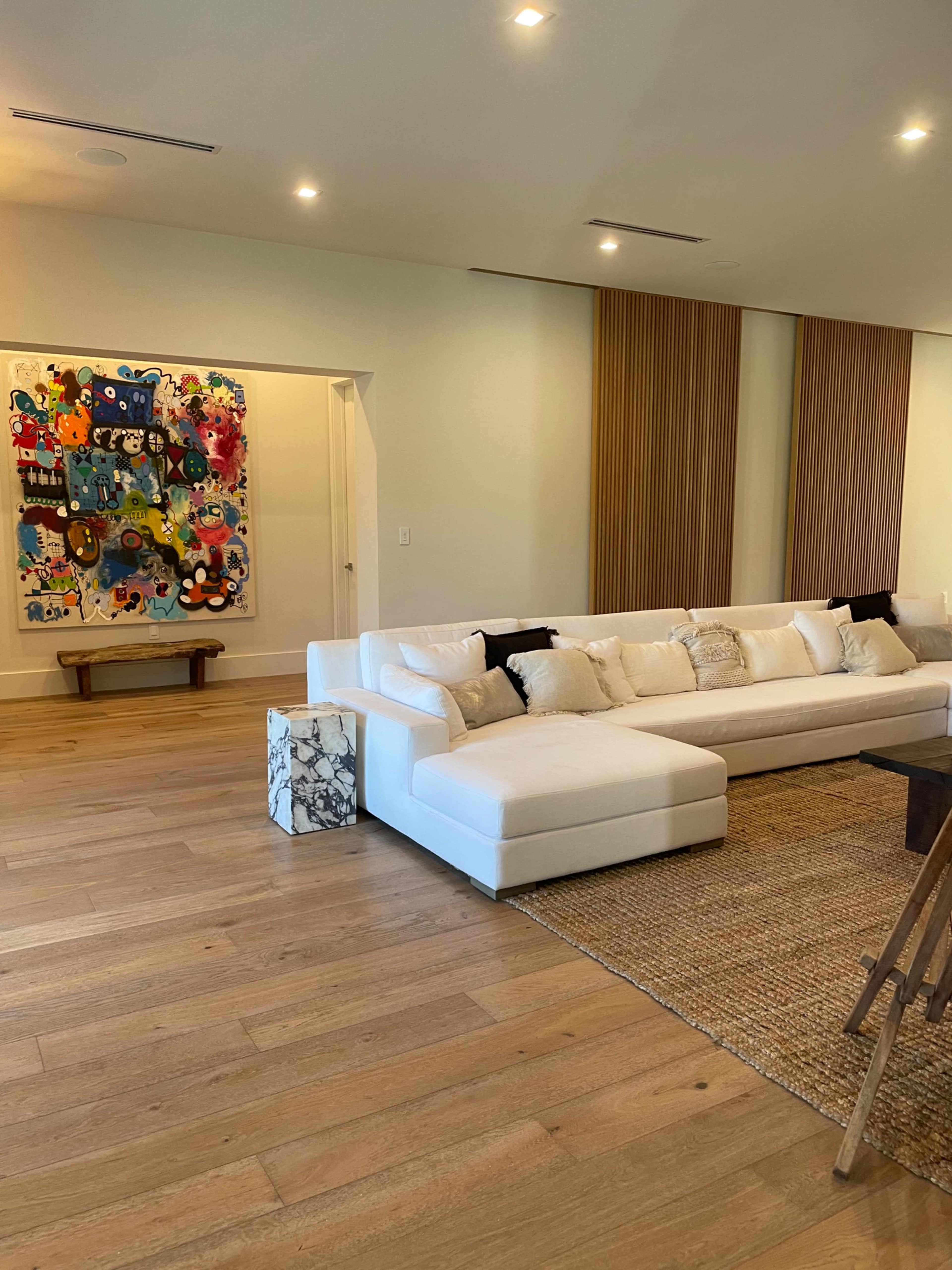 The image shows a spacious living room featuring a large white sectional sofa, a textured area rug, and a colorful artwork hung on the wall.