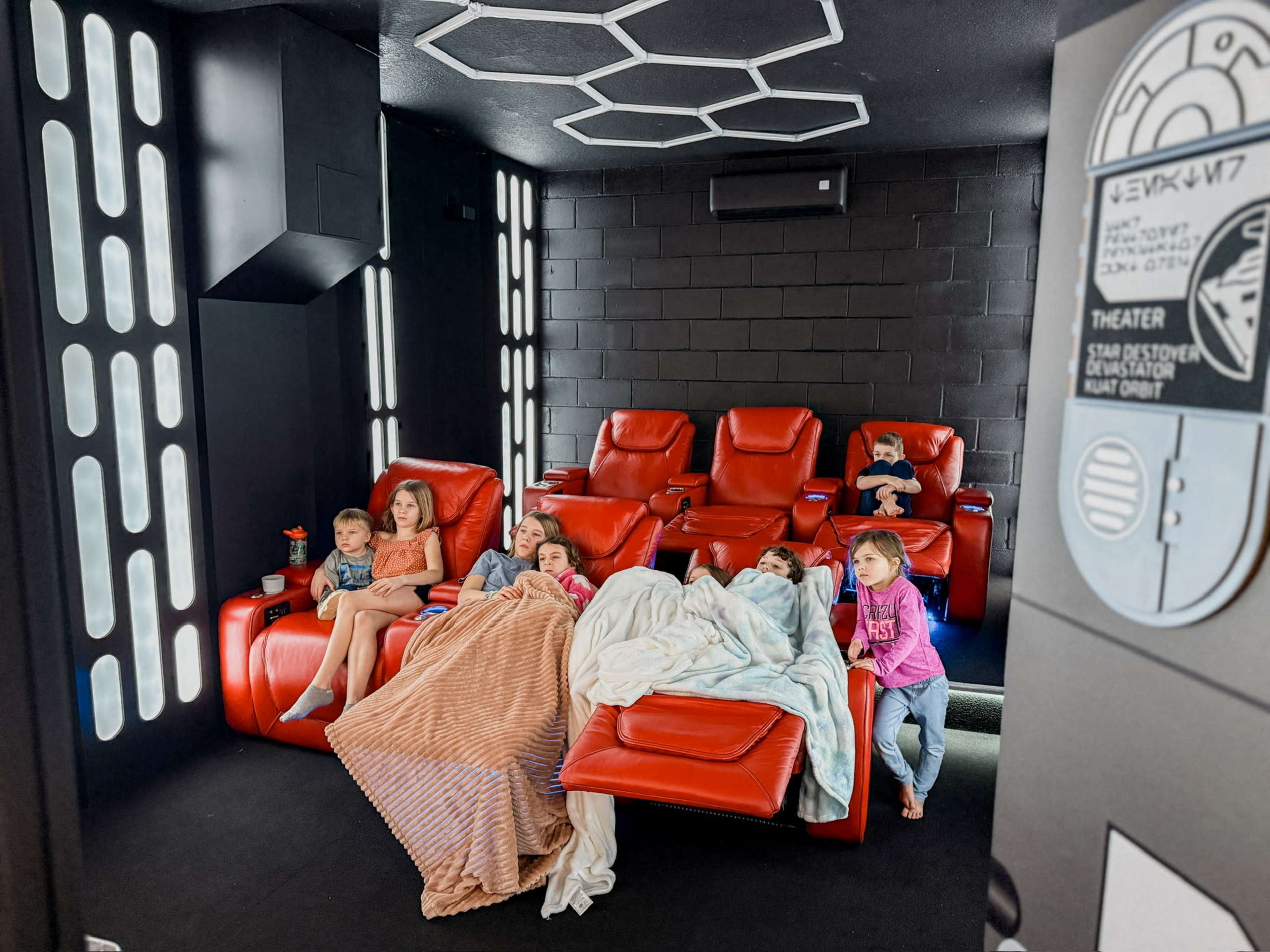 A group of children sits on red recliners in a home theater, watching a movie while wrapped in blankets.