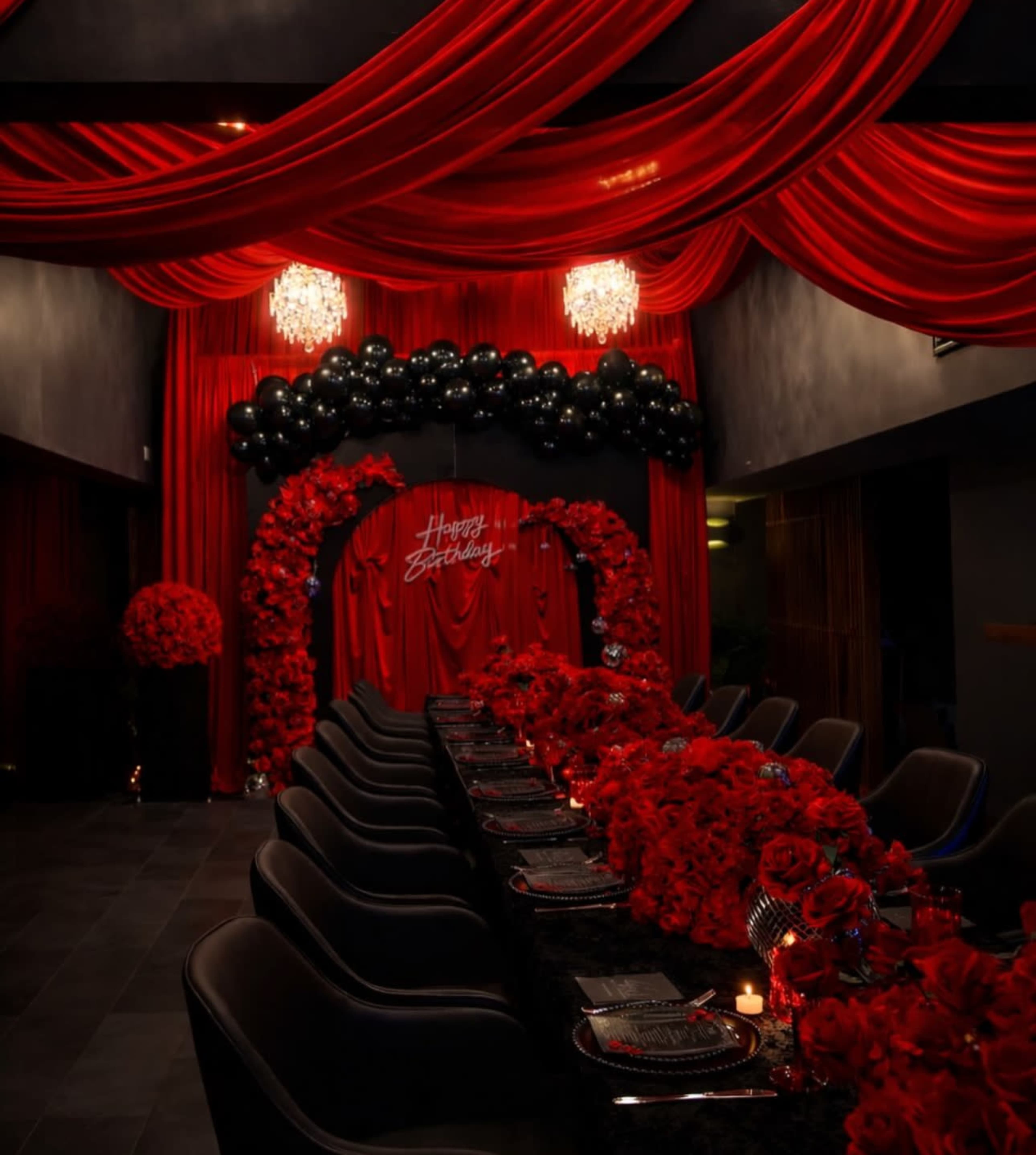 The image depicts a lavish birthday celebration venue adorned with red drapery, black balloons, and an abundance of red flowers.