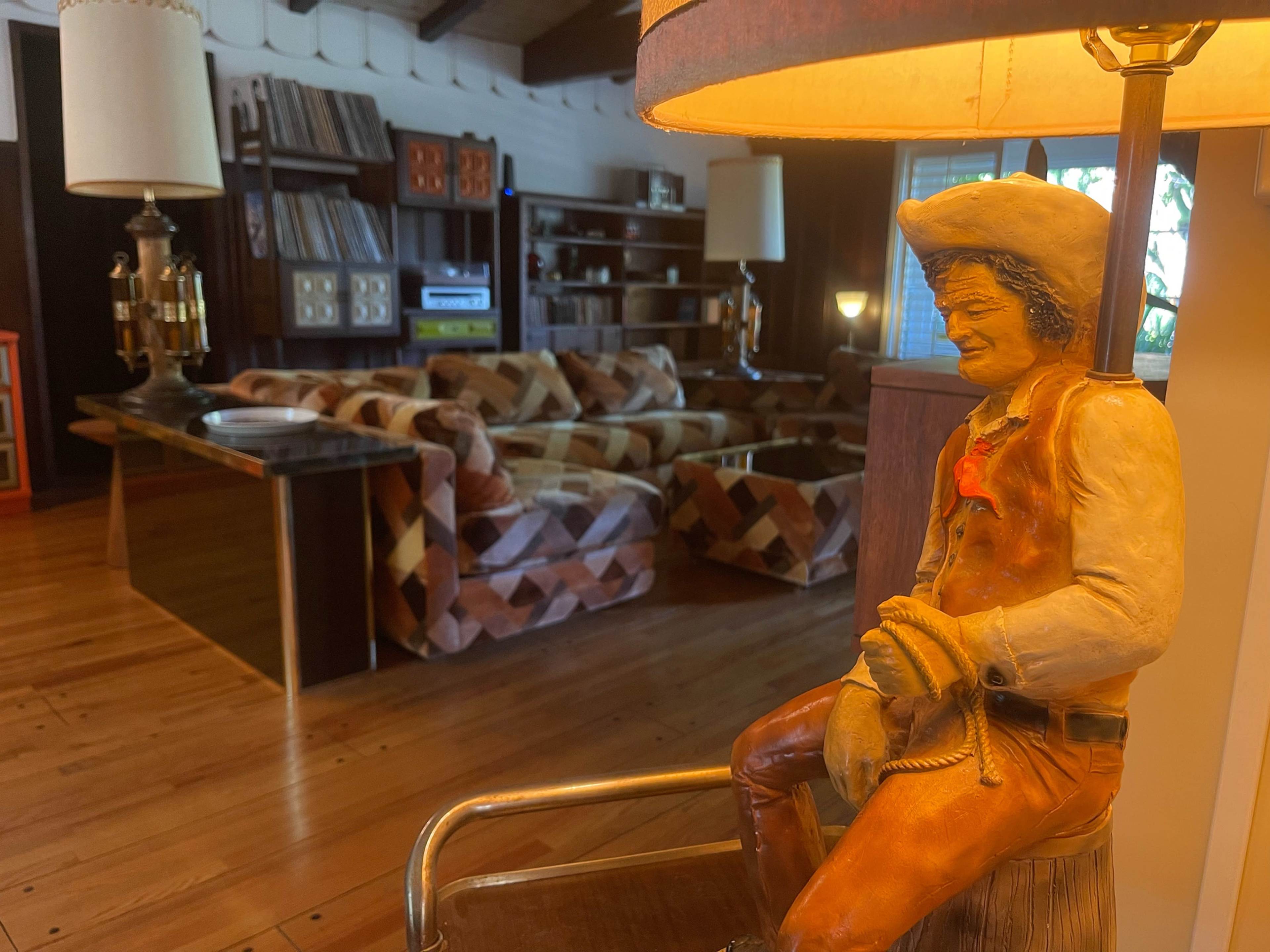 A cowboy figurine sits on a table in a room featuring a patterned sofa, wooden flooring, and shelves filled with records.
