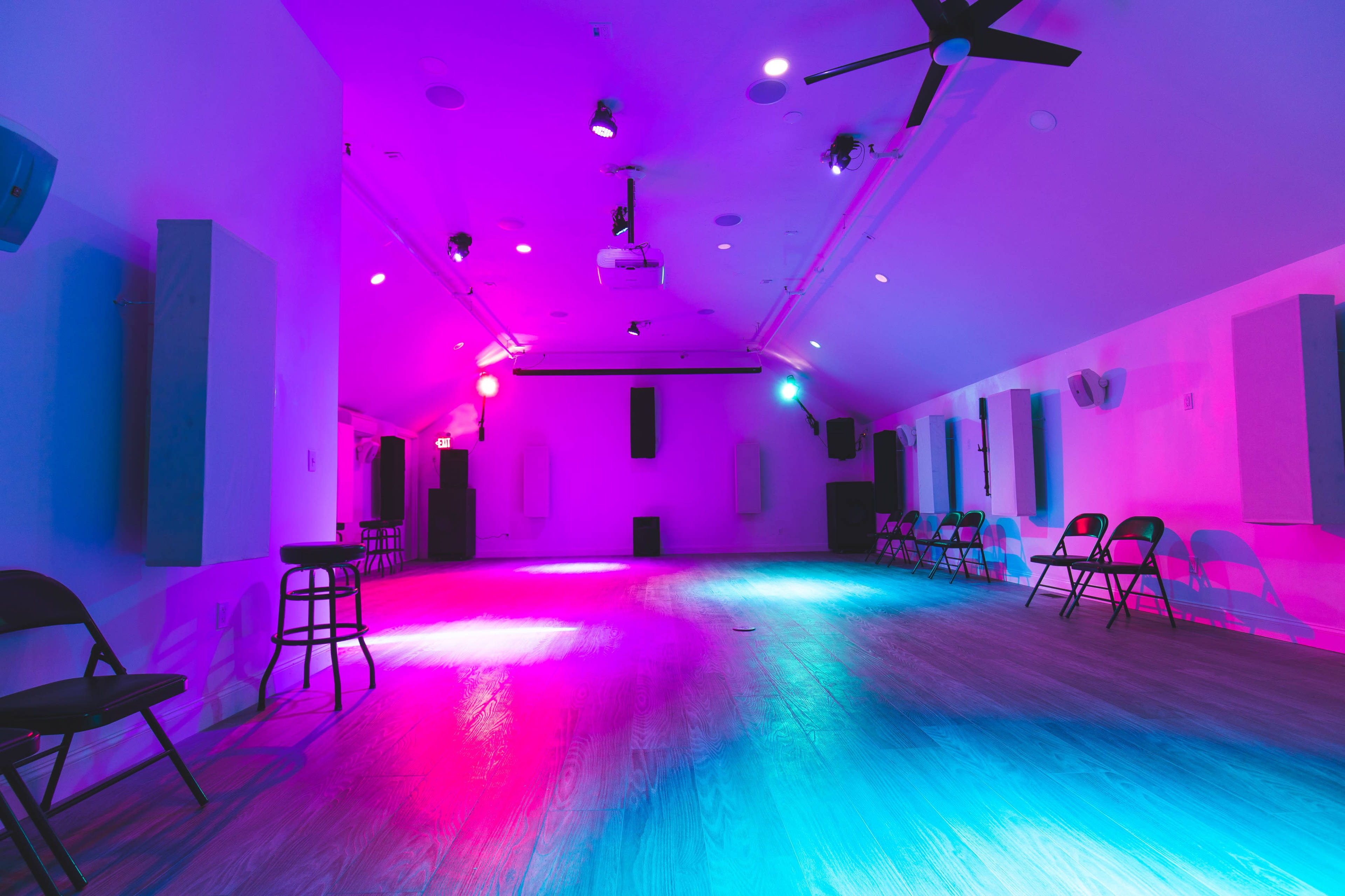 A spacious room with wooden flooring is illuminated by purple and blue lights, featuring rows of black folding chairs along the sides.