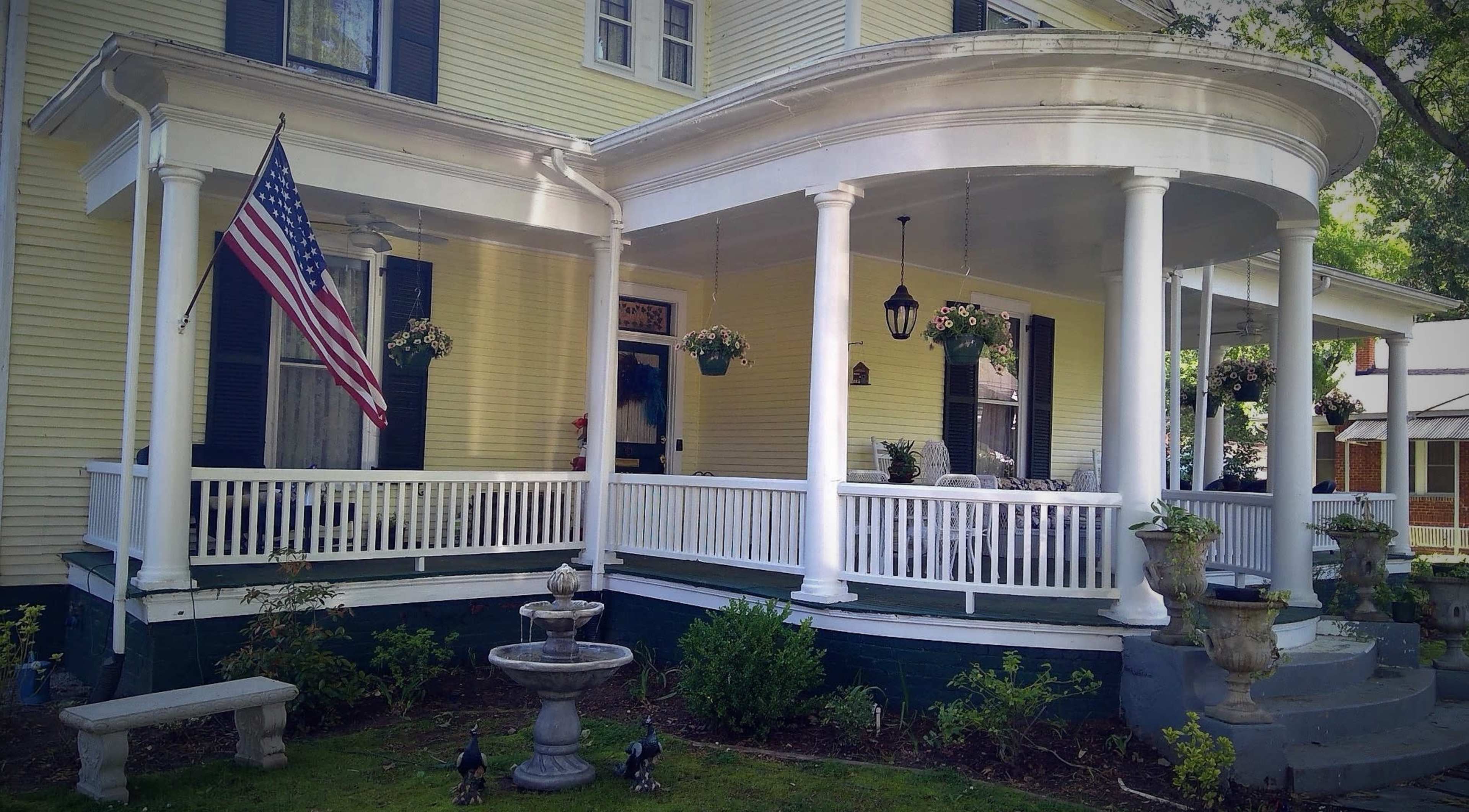 The image shows a yellow house with a wraparound porch, American flag, potted plants, and a decorative fountain in the front yard.