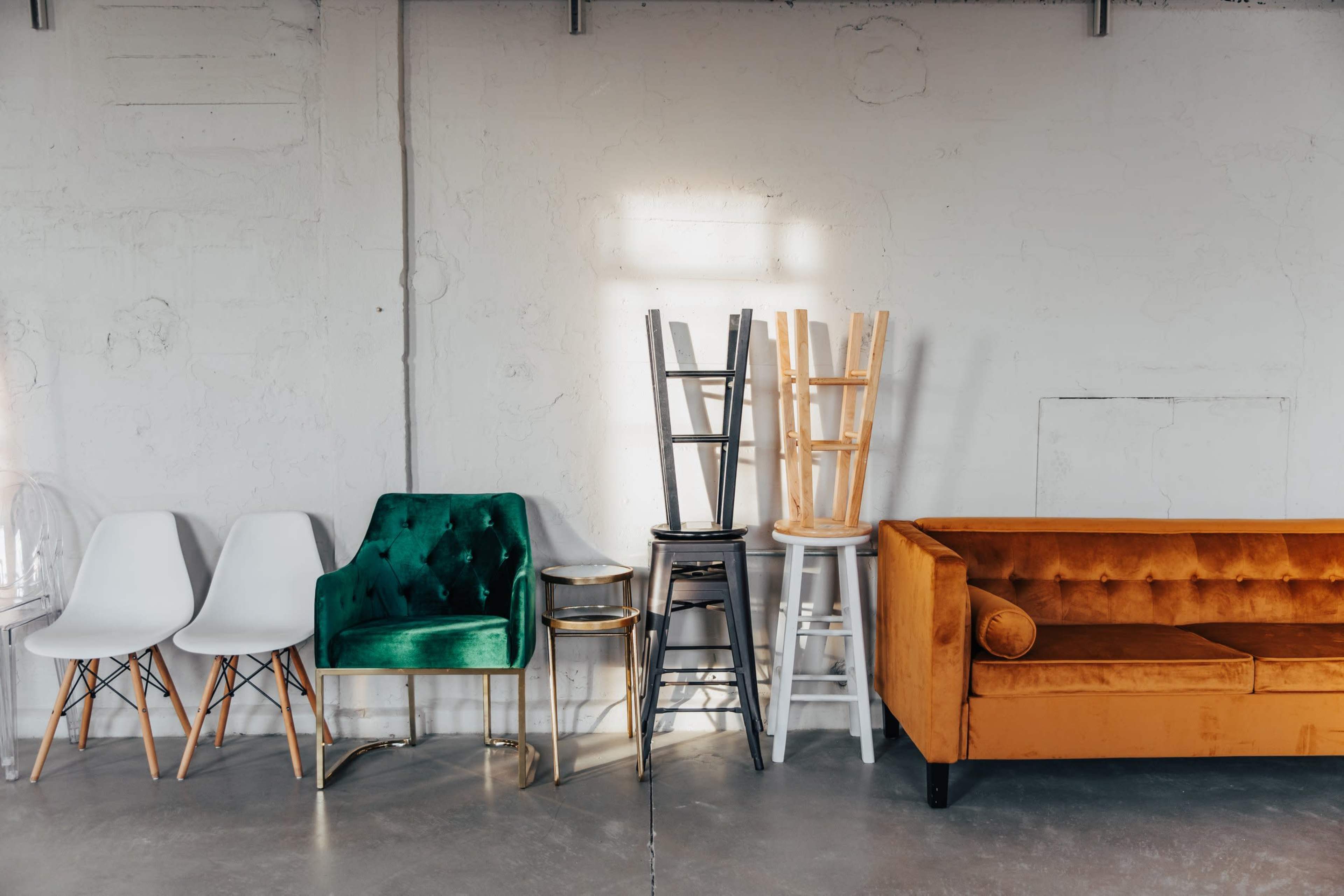 A modern interior scene with a green velvet chair, white and metallic stools stacked on a table, and a tufted orange couch against a concrete wall.