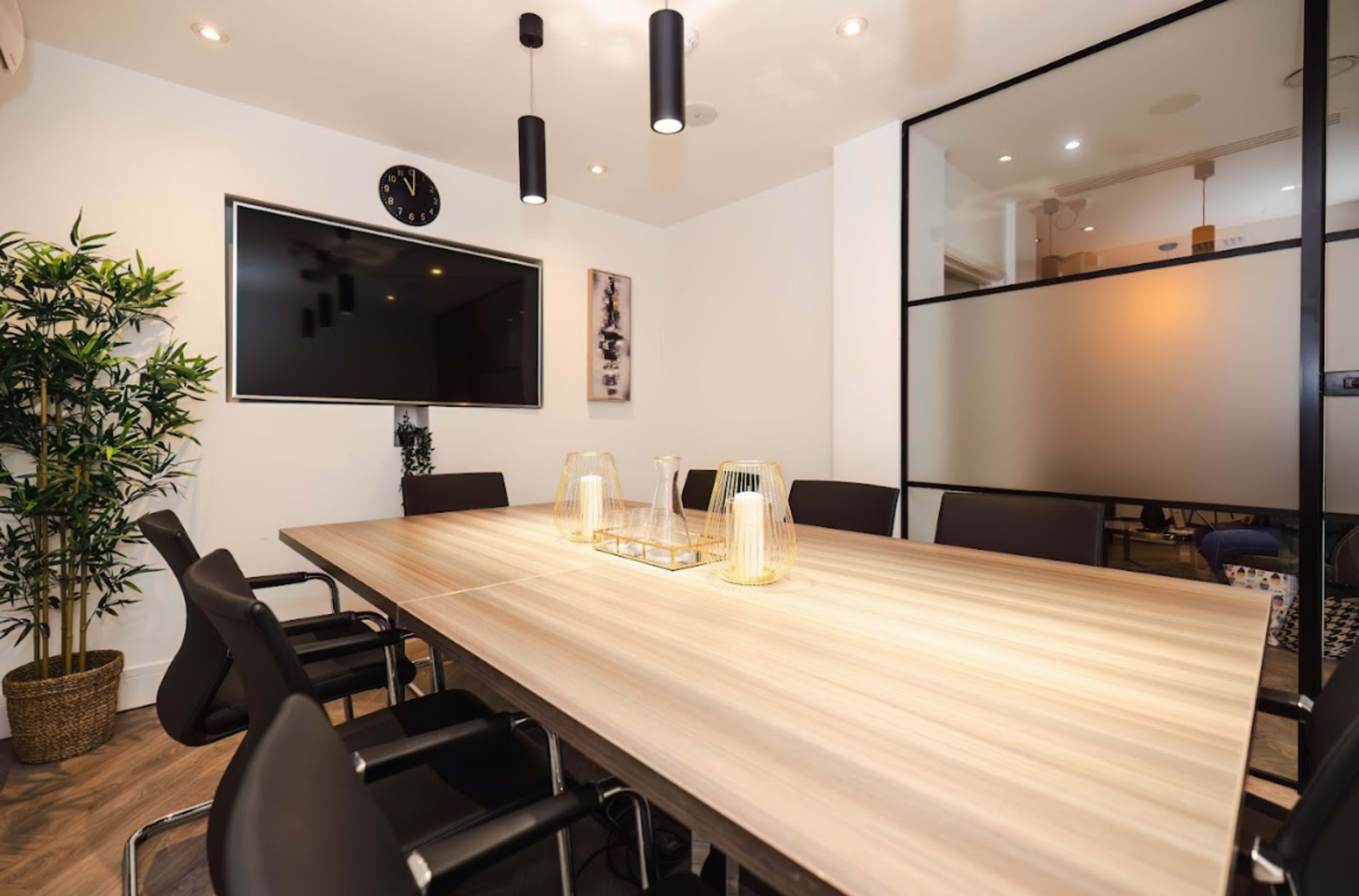 A modern conference room features a large wooden table surrounded by black chairs, with a television mounted on the wall and decorative plants.