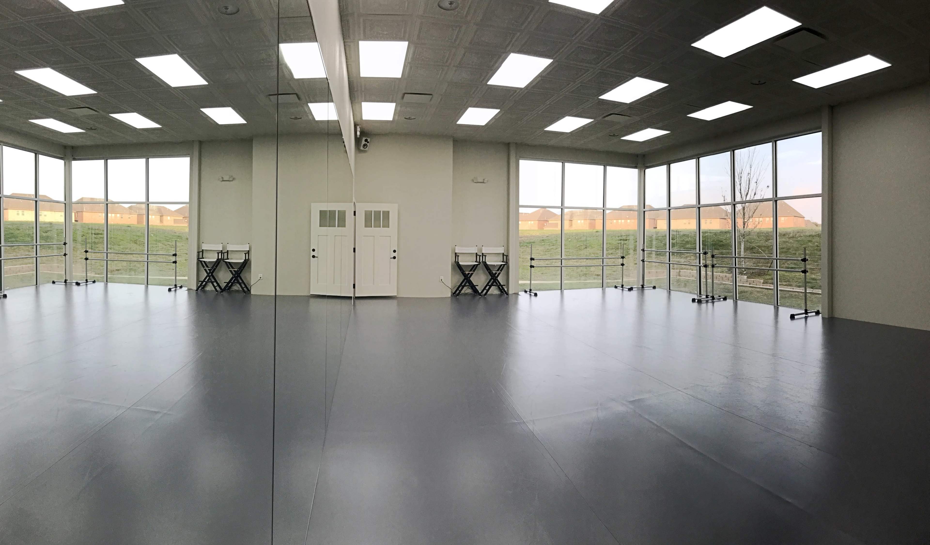 A spacious, empty room with large windows, white walls, and a gray floor, featuring tables on one side and double doors on the opposite side.