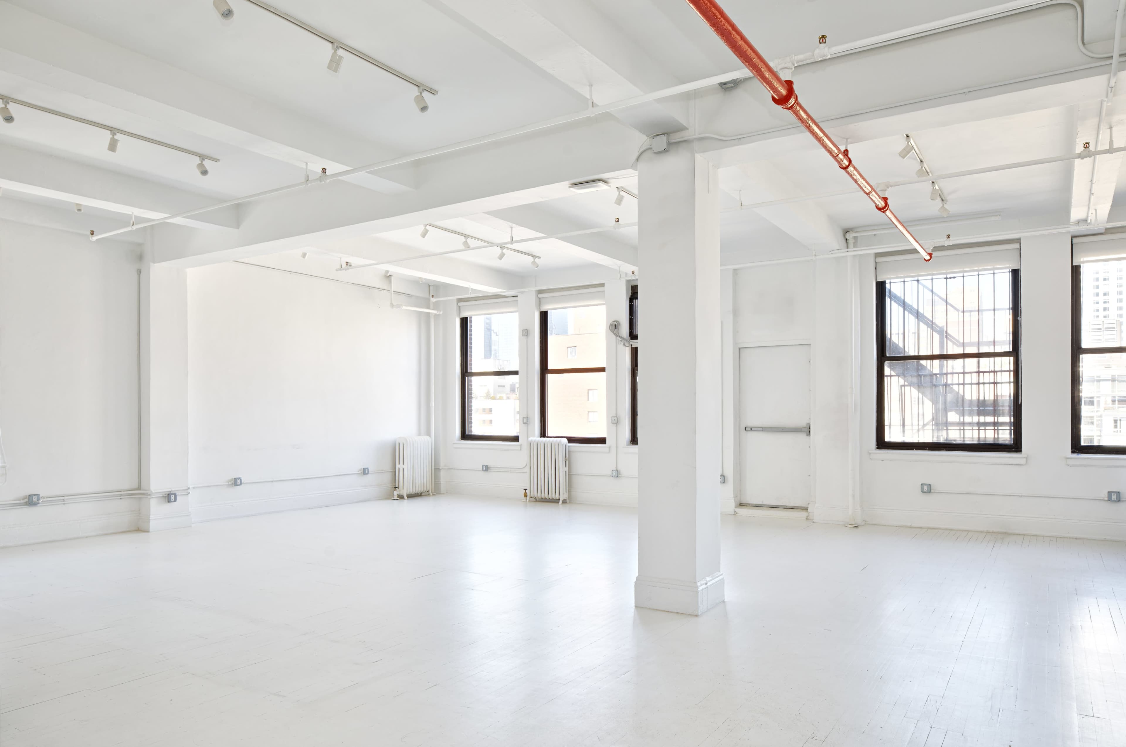 A spacious, empty room with white walls, large windows, and bright overhead lighting.