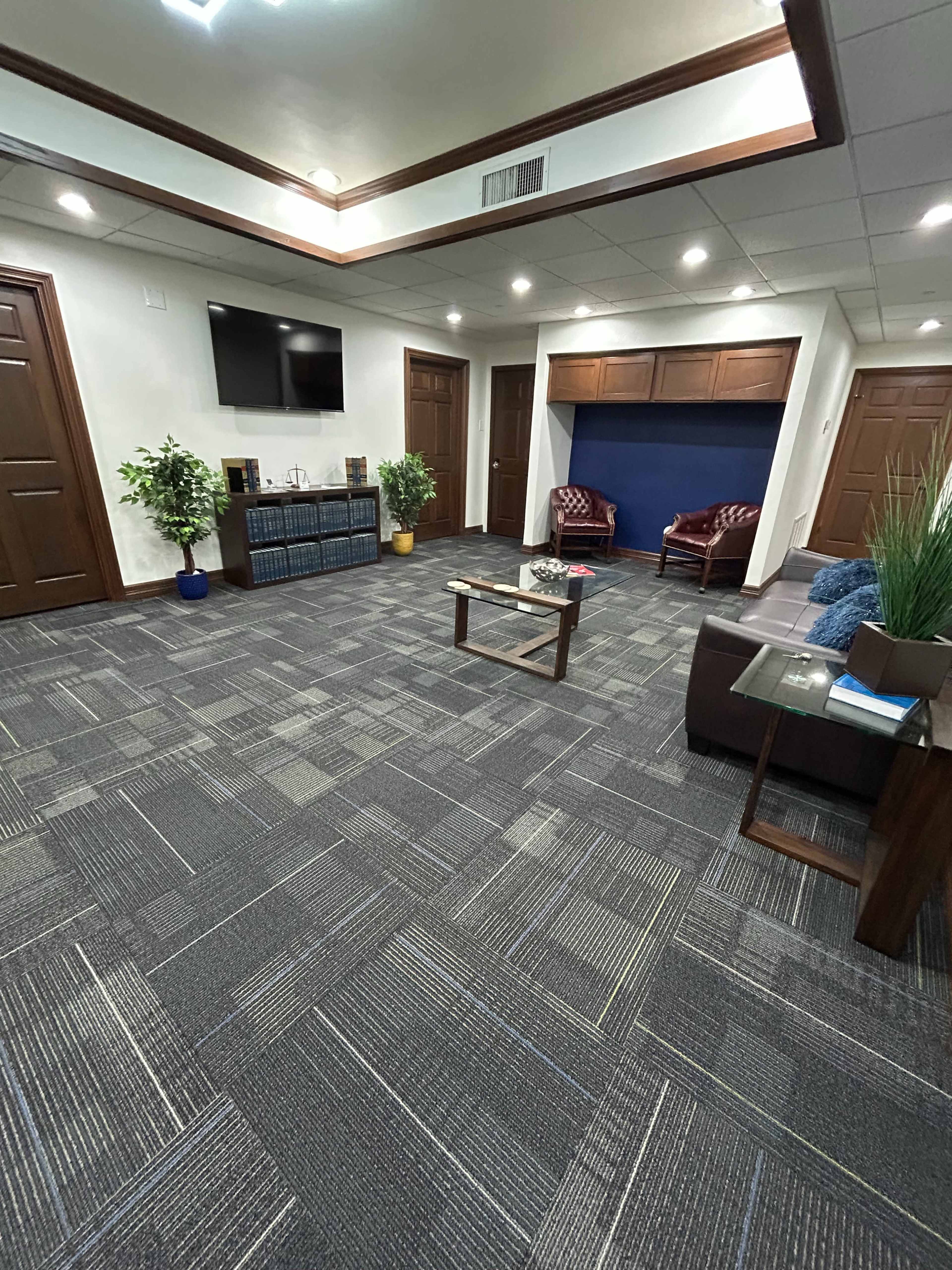 The image shows a spacious waiting area with two armchairs, a coffee table, plants, and a wall-mounted television.