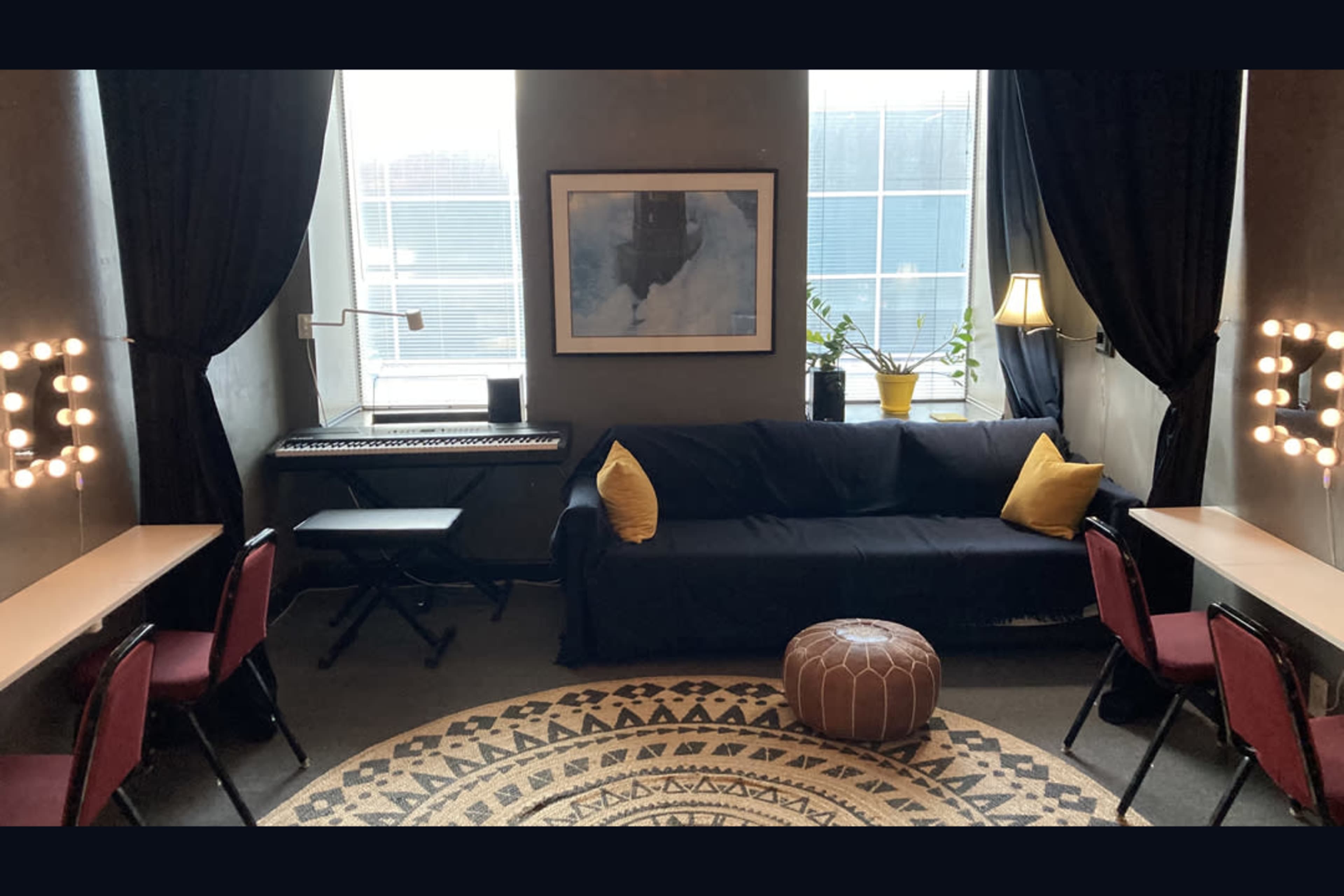 The image shows a cozy room with a black couch, a keyboard, two small tables with chairs, and a patterned rug, all set against a backdrop of large windows and soft lighting.