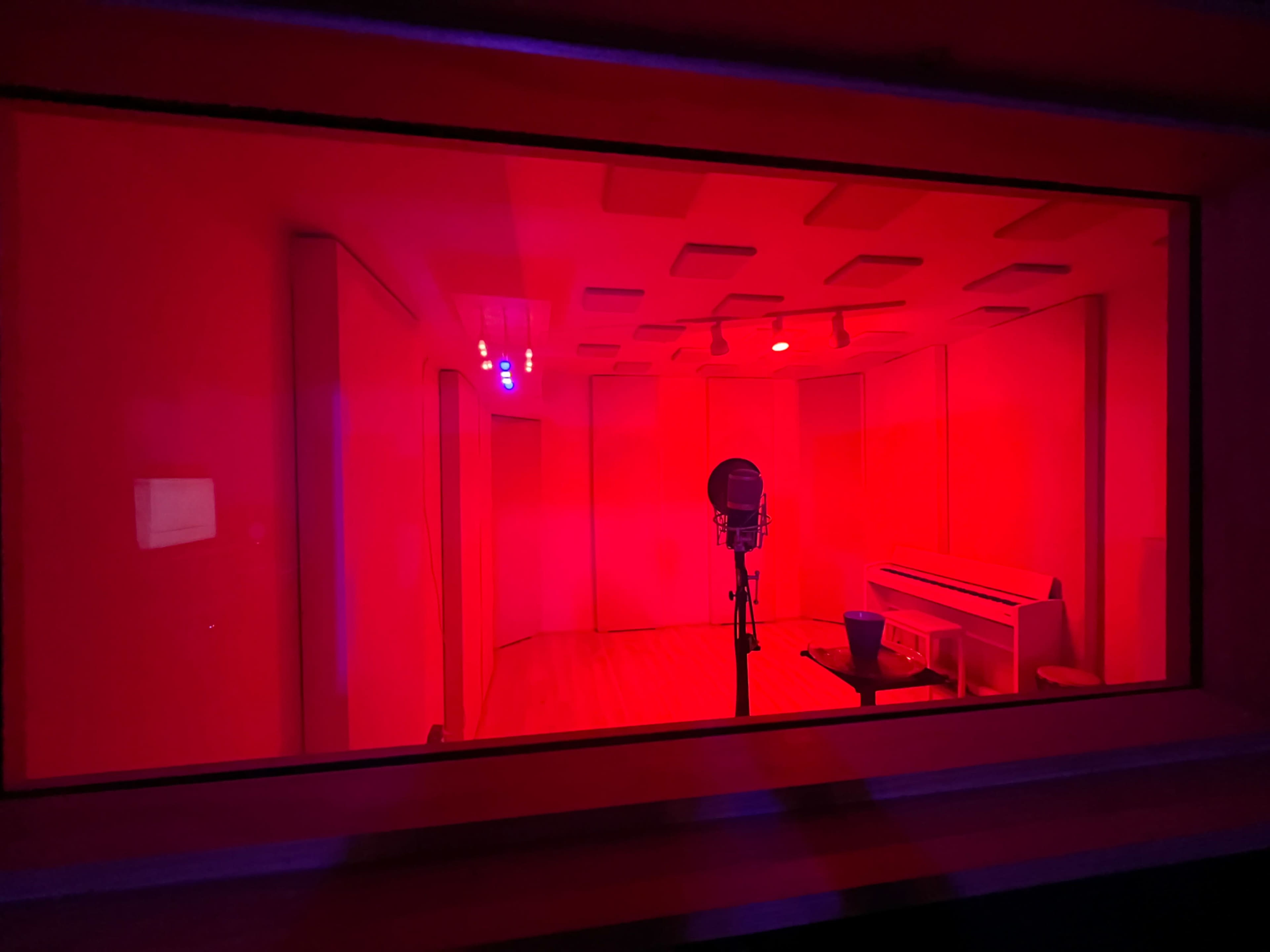 The image shows a recording studio with red lighting, featuring a microphone positioned in the center and soundproof panels on the walls.