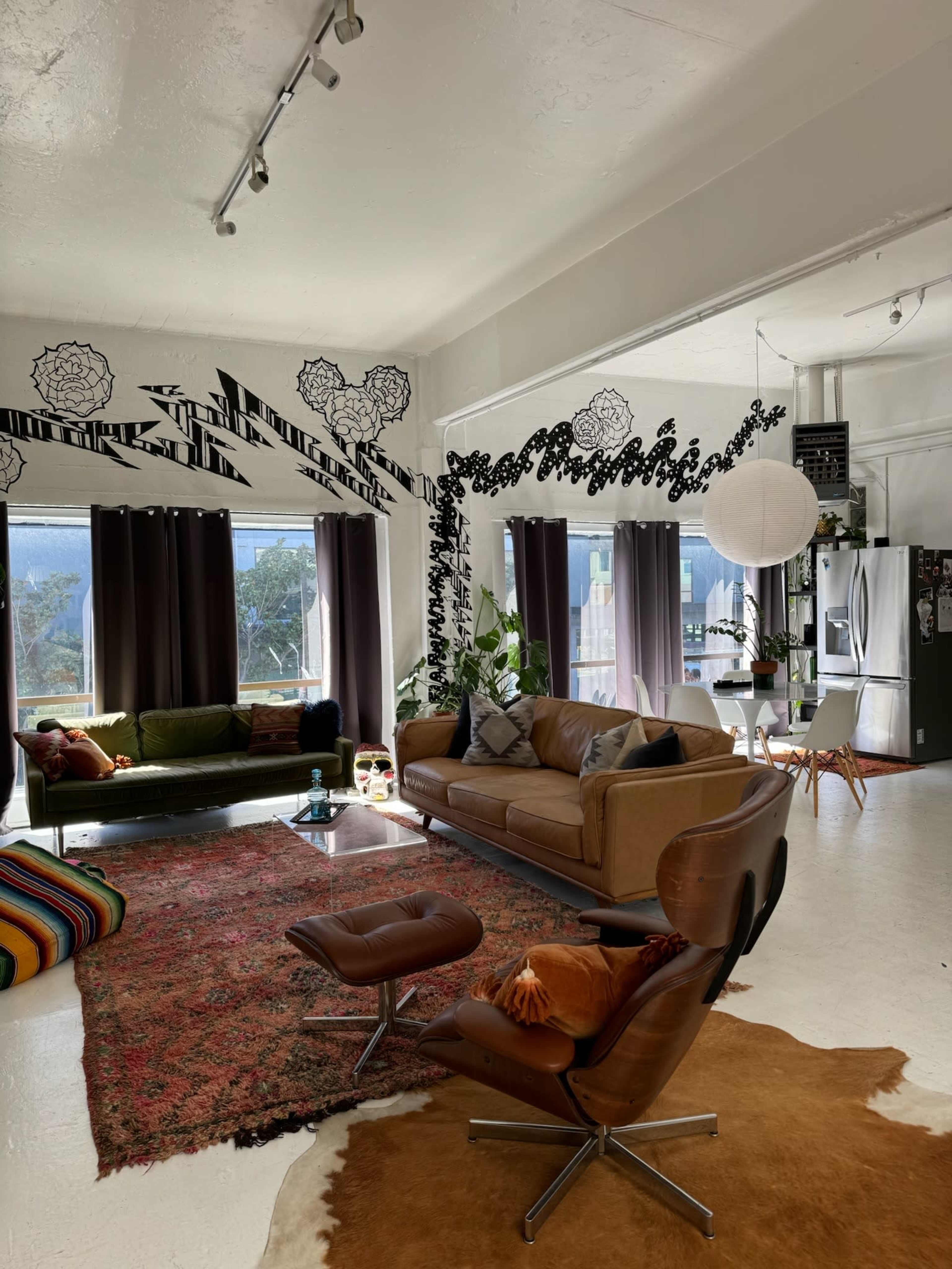 The image depicts a stylish living room with a mix of modern furniture, large windows allowing natural light, and decorative wall patterns.