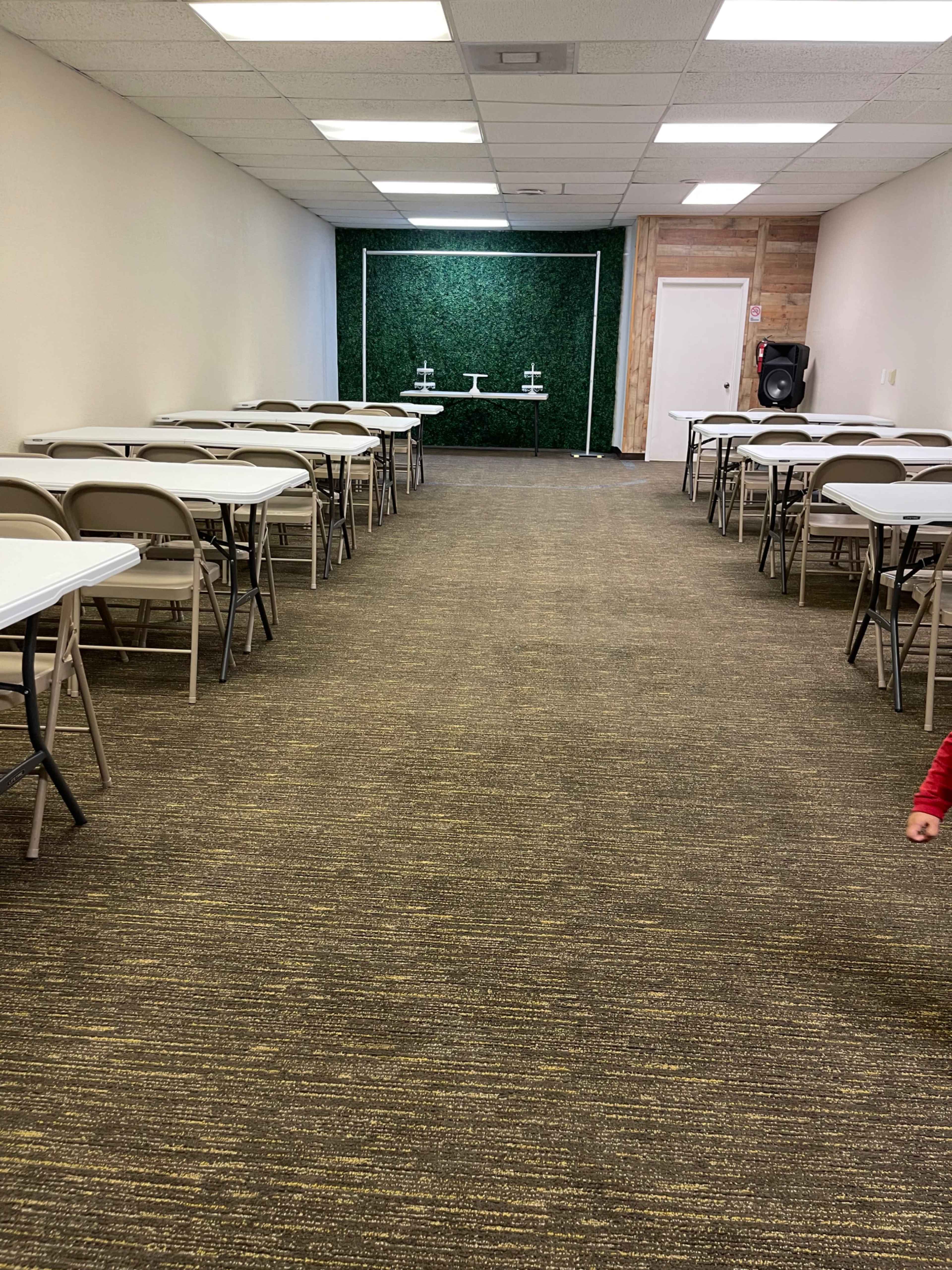 A narrow room with rows of tables and chairs on both sides, leading to a green wall with two white display stands at the far end.
