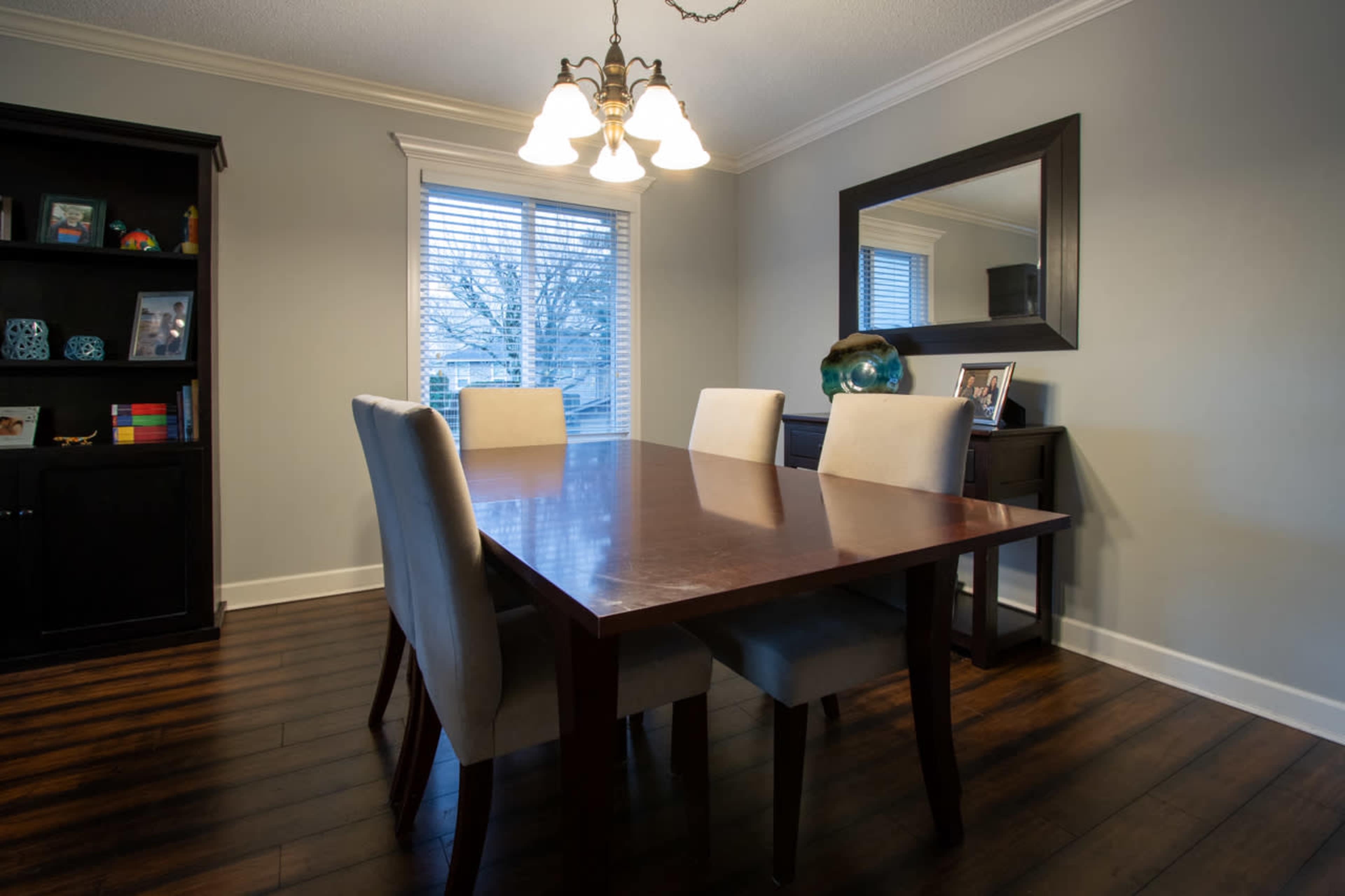 A dining room features a rectangular wooden table surrounded by four upholstered chairs, with a black bookshelf and a large mirror on the walls.