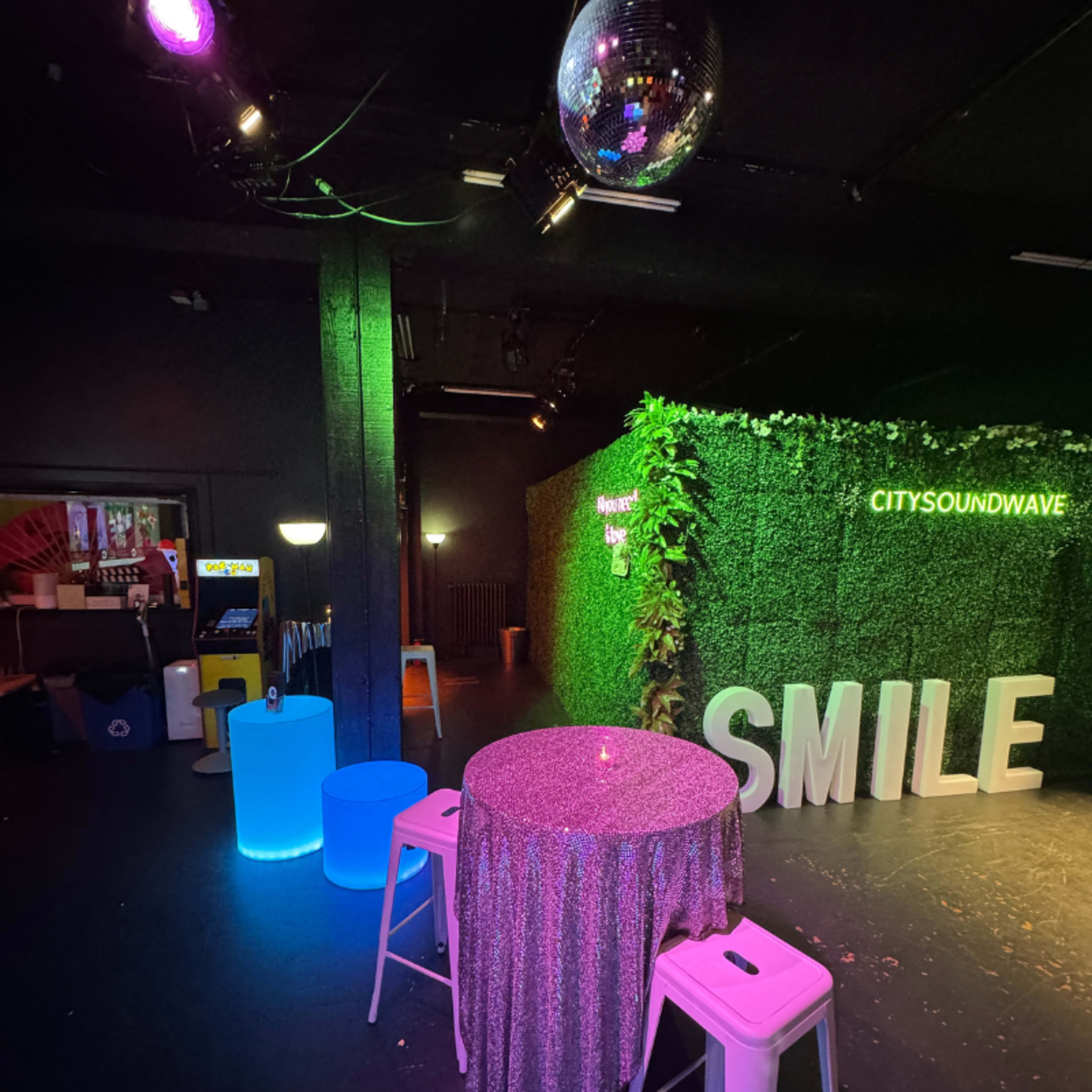 The image shows a dimly lit venue featuring a green wall with the word "SMILE" and a bar area with colorful illuminated furniture.