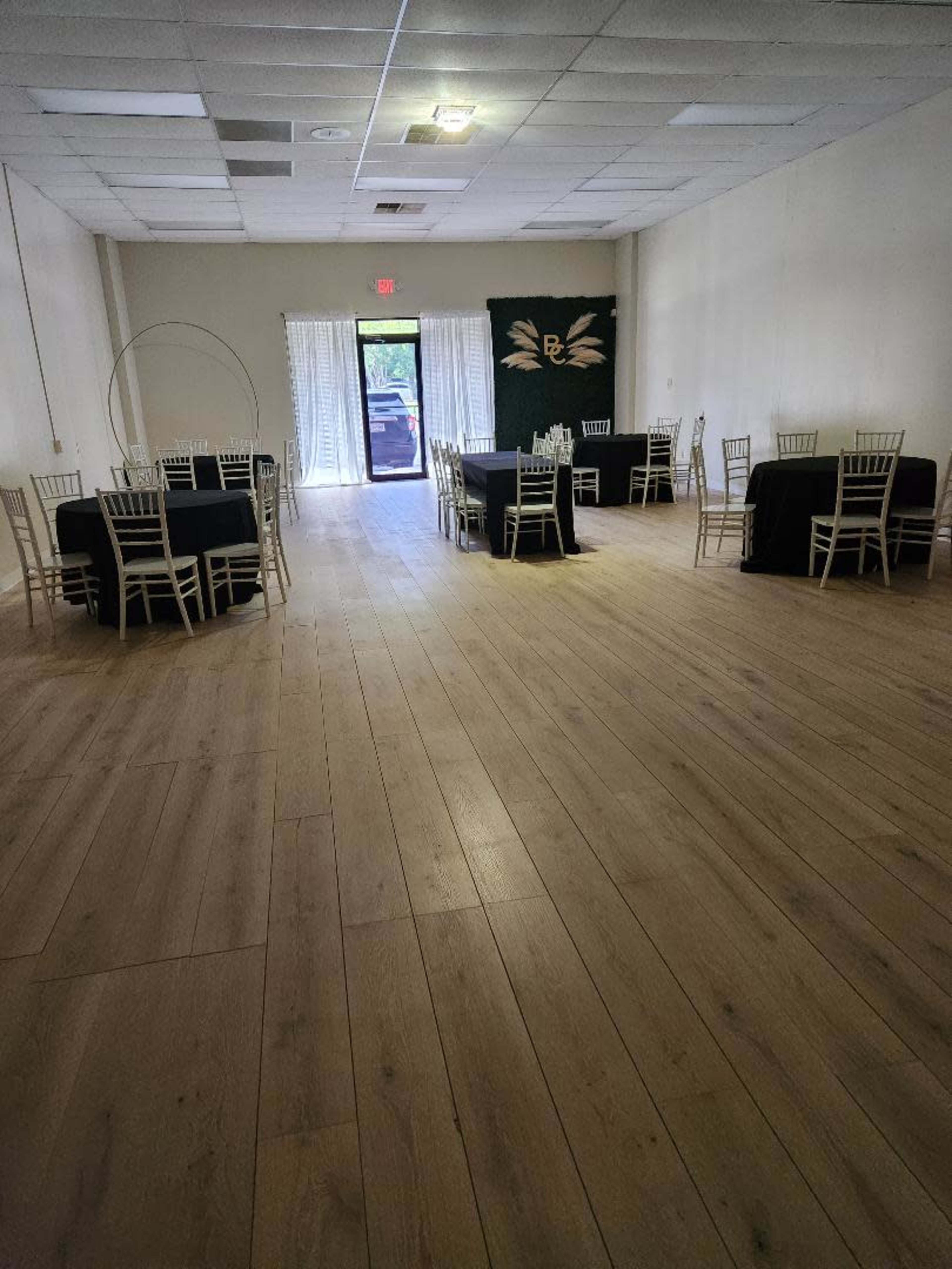 A spacious event room with several round tables covered in black cloth and wooden flooring, leading to a doorway at the back.
