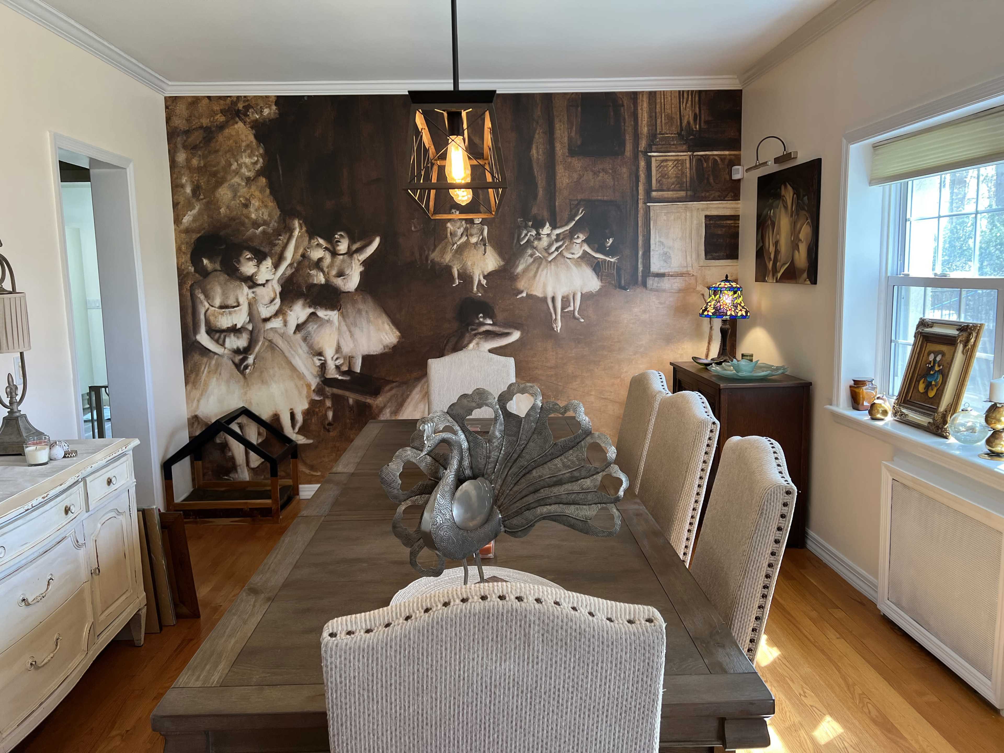 A dining room features a wooden table surrounded by upholstered chairs, with a large mural of ballet dancers on one wall and a decorative peacock sculpture at the center.