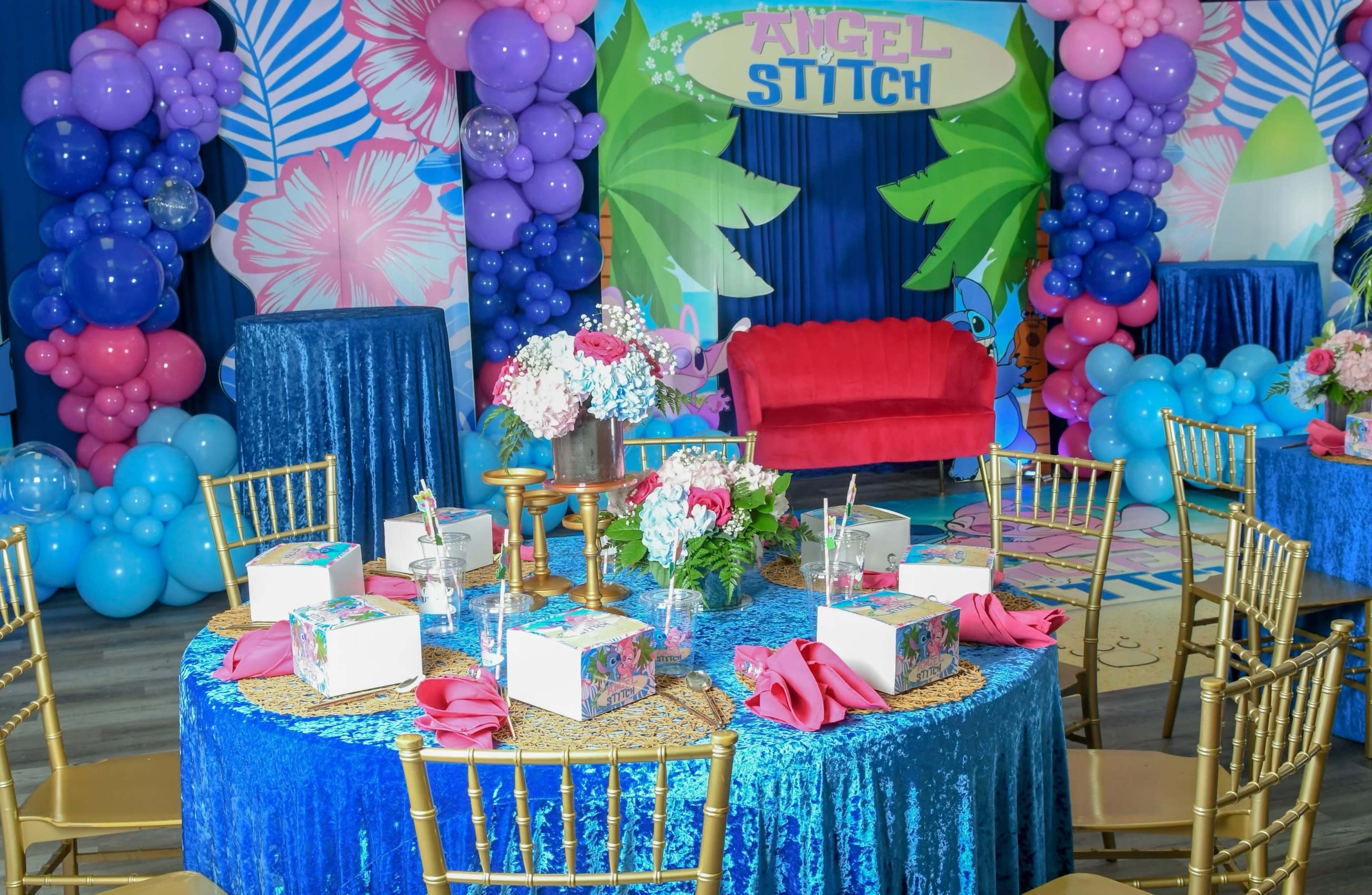 The scene features a decorated party space with tables dressed in blue and adorned with floral centerpieces, surrounded by colorful balloons and a backdrop displaying "Angel Stitch."