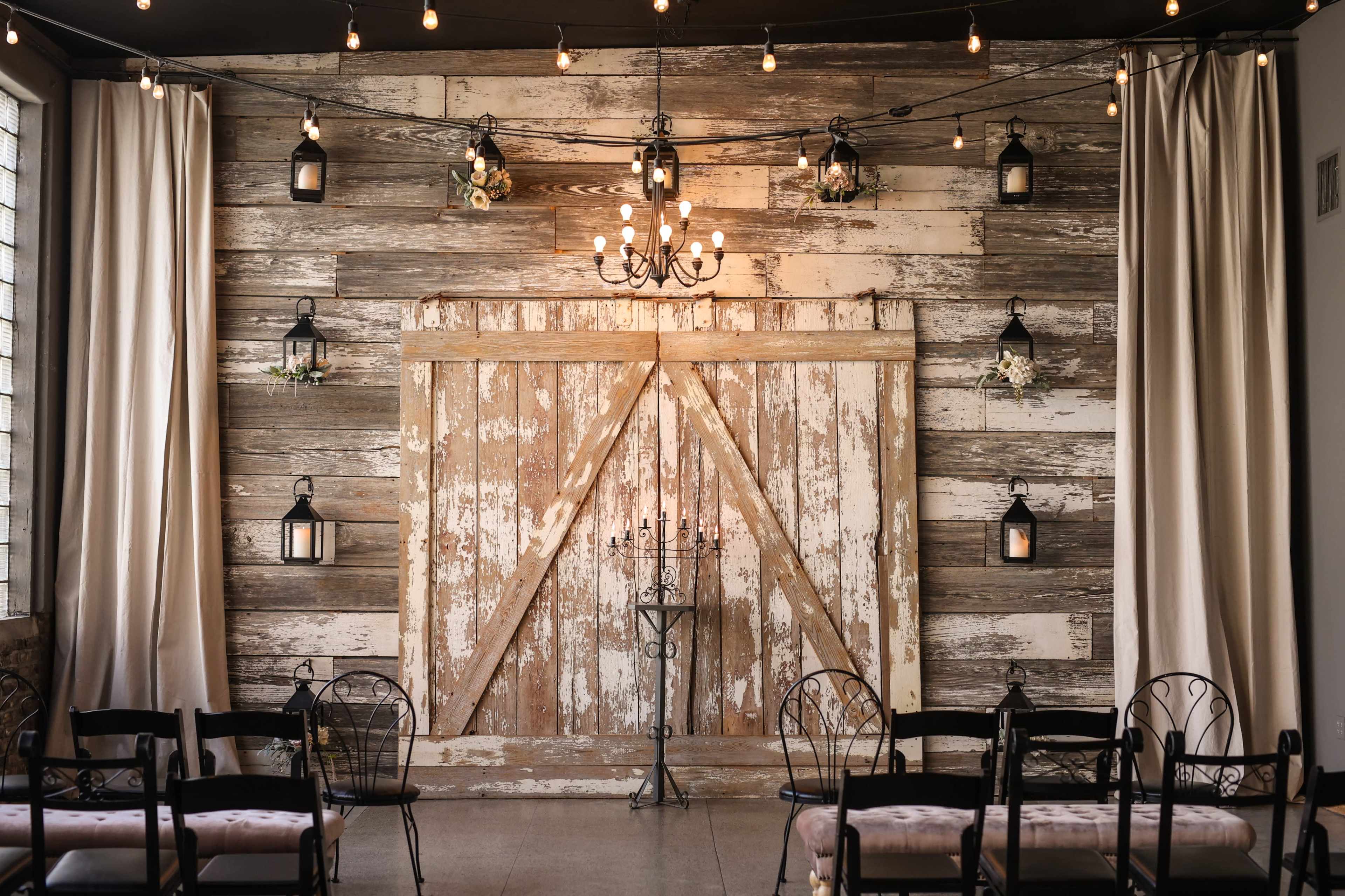A rustic interior space with a weathered wooden wall and a large barn door, illuminated by hanging lights and featuring decorative lanterns.