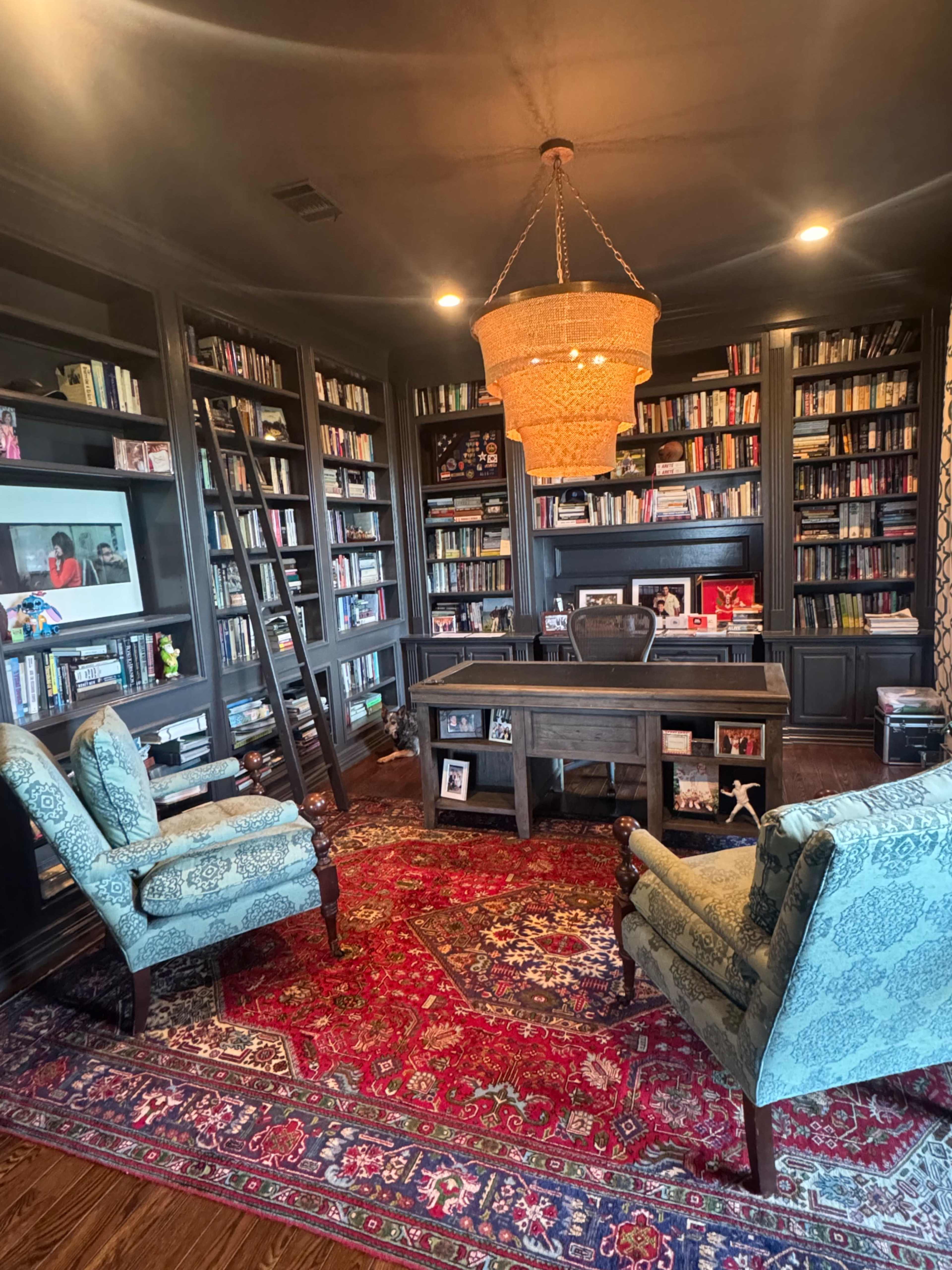 The image depicts a cozy home office with a large bookshelf filled with books, a wooden desk at the center, and two upholstered chairs on a patterned rug beneath a chandelier.