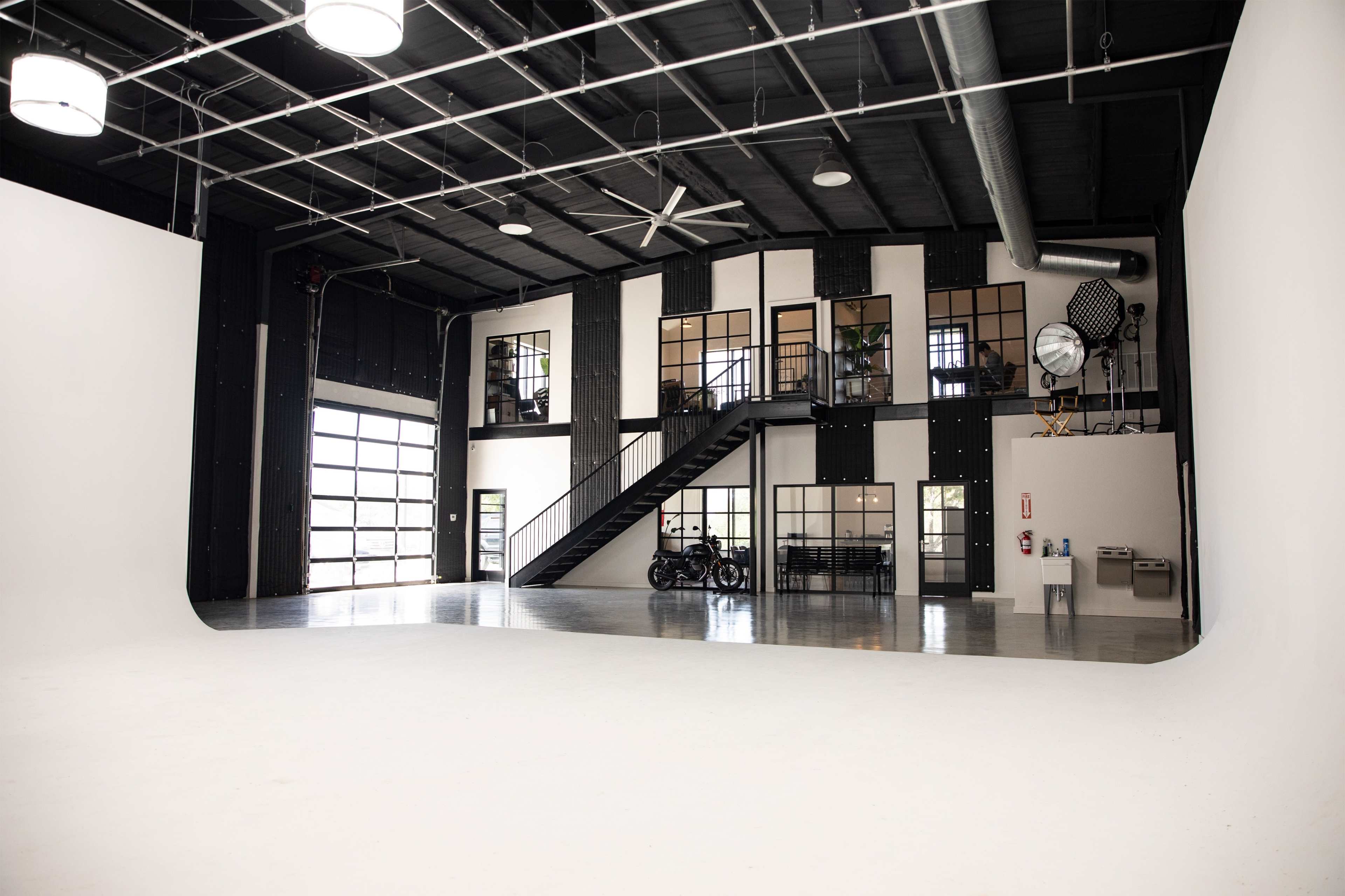 A large, well-lit studio space with a white curved backdrop, high ceilings, and a staircase leading to a mezzanine level with large windows.