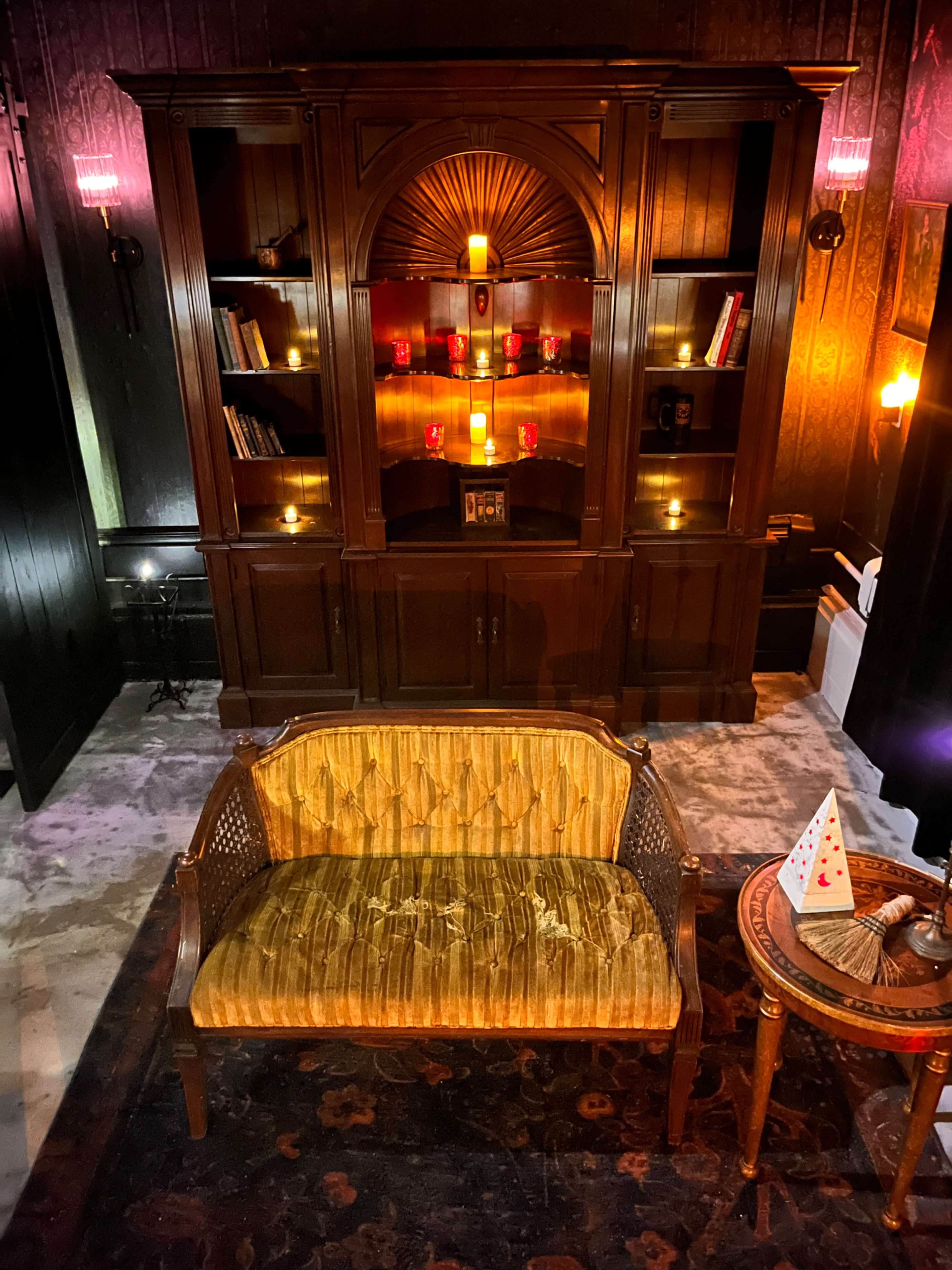 The image depicts a cozy room featuring a wooden cabinet with shelves illuminated by candles, a vintage seating area with a patterned sofa, and a small round table with a decorative item.