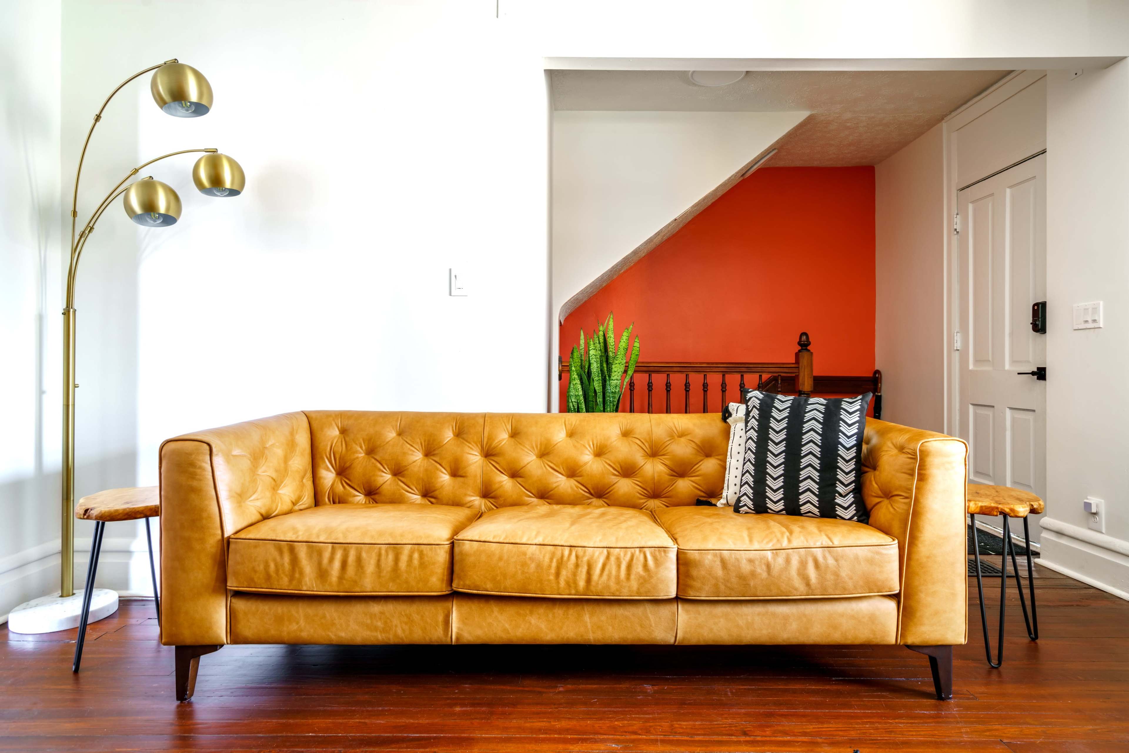 A tufted, tan leather sofa with decorative pillows sits in a well-lit room, with a tall floor lamp and a staircase in the background.