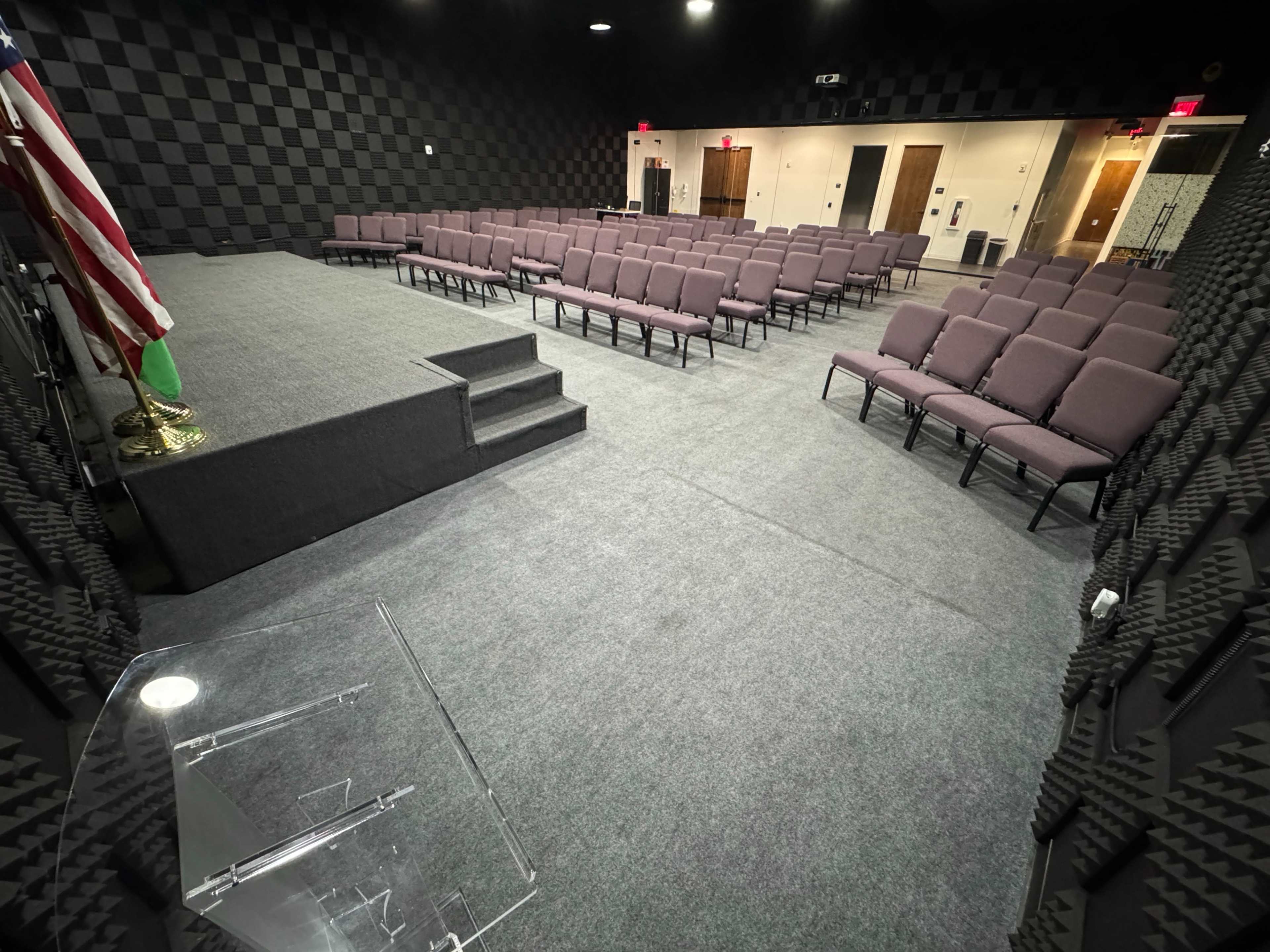 The image shows a small auditorium with seating arranged in rows facing a stage, featuring a podium and an American flag on display.