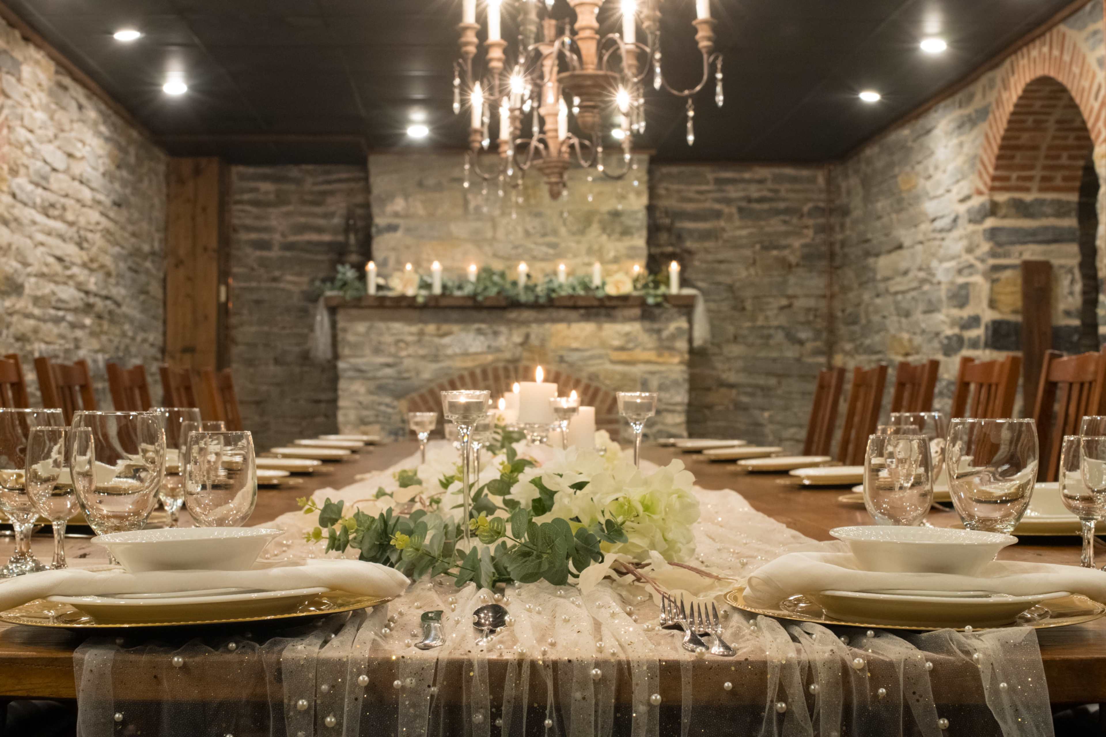 A long dining table is set with elegant dinnerware, surrounded by chairs, in a stone-walled room with soft candlelight illuminating the space.