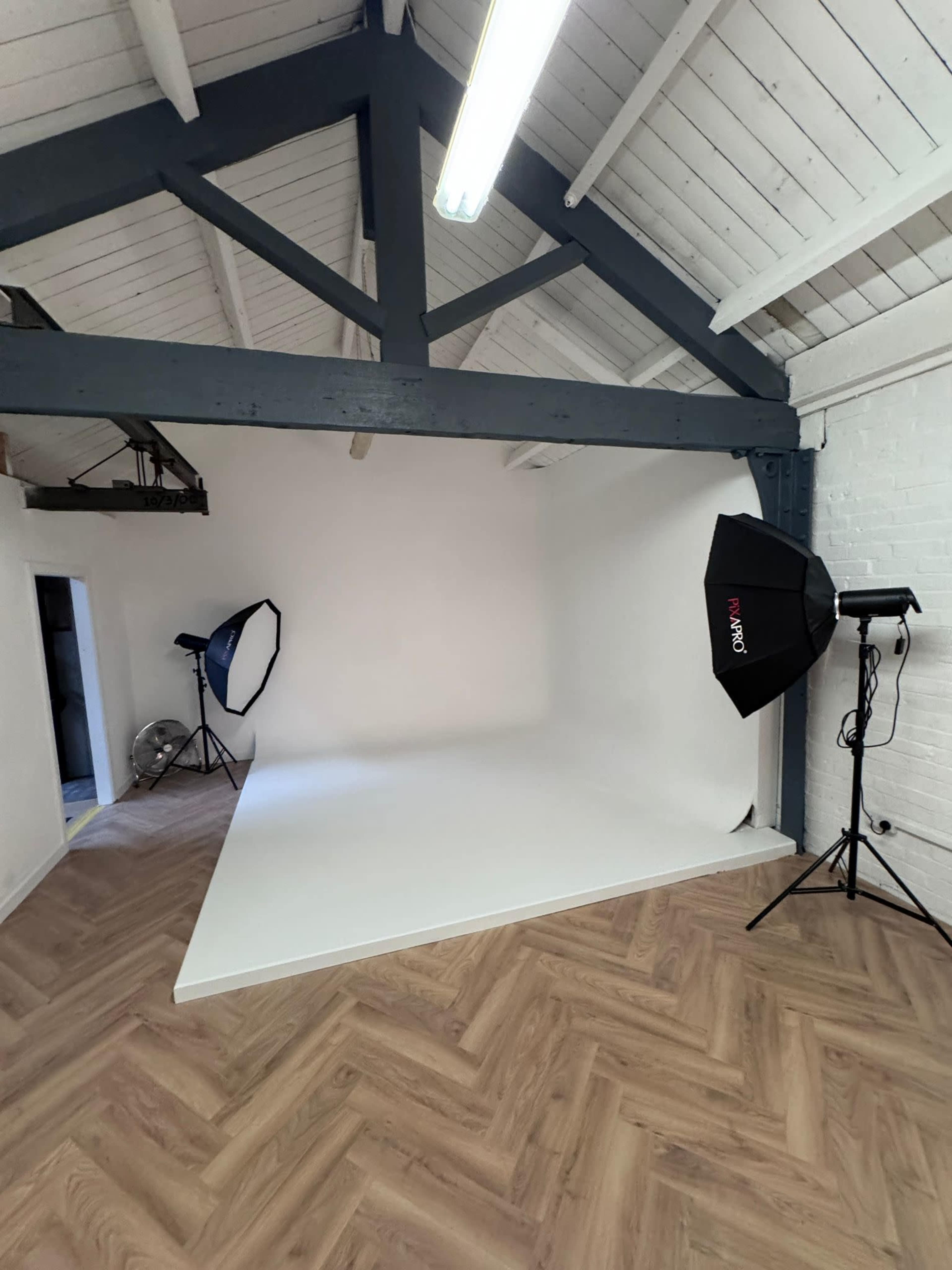 The image shows a photography studio with a white backdrop, two softbox lights, and wooden flooring.