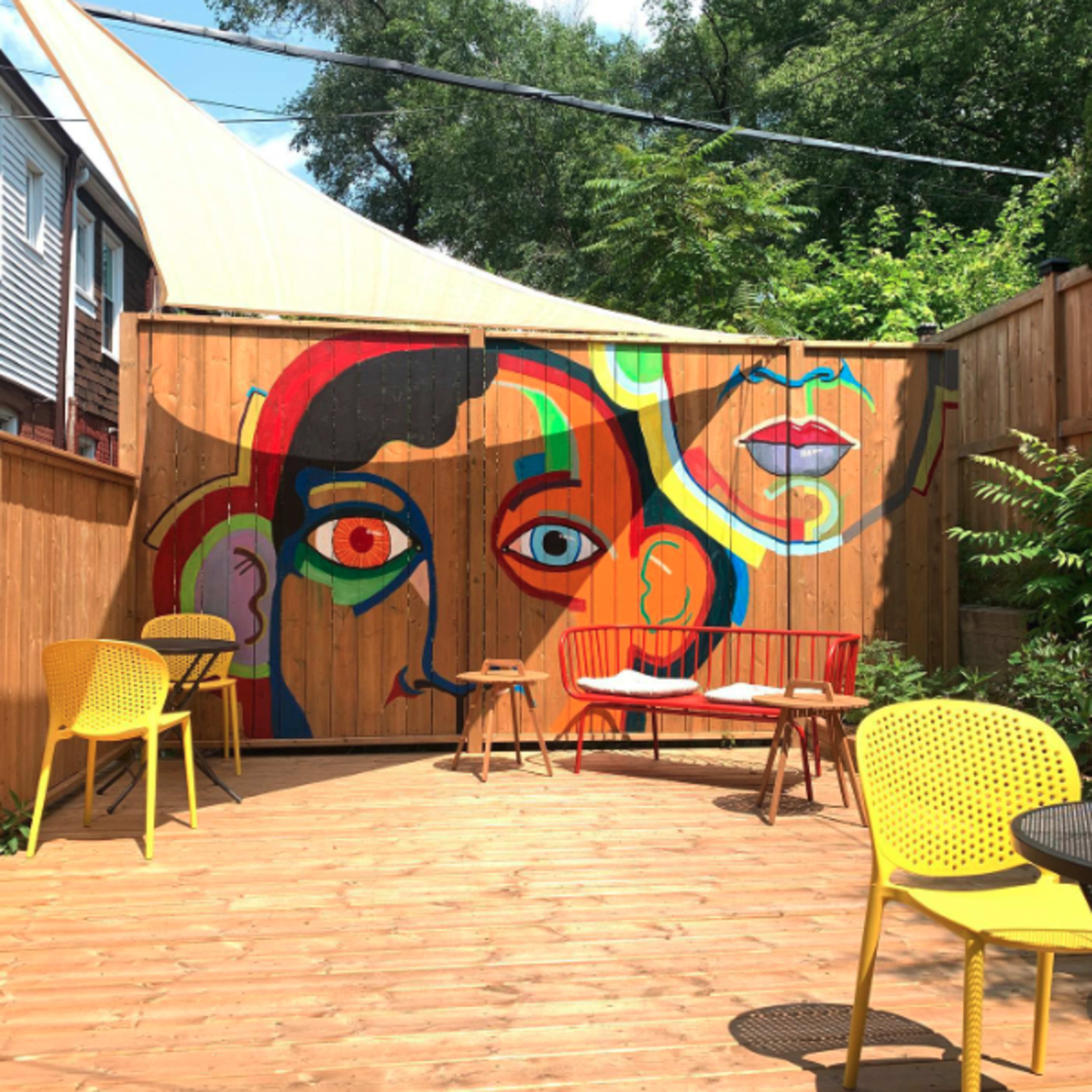 A wooden patio area with colorful chairs and a large mural depicting abstract faces on a wooden fence.