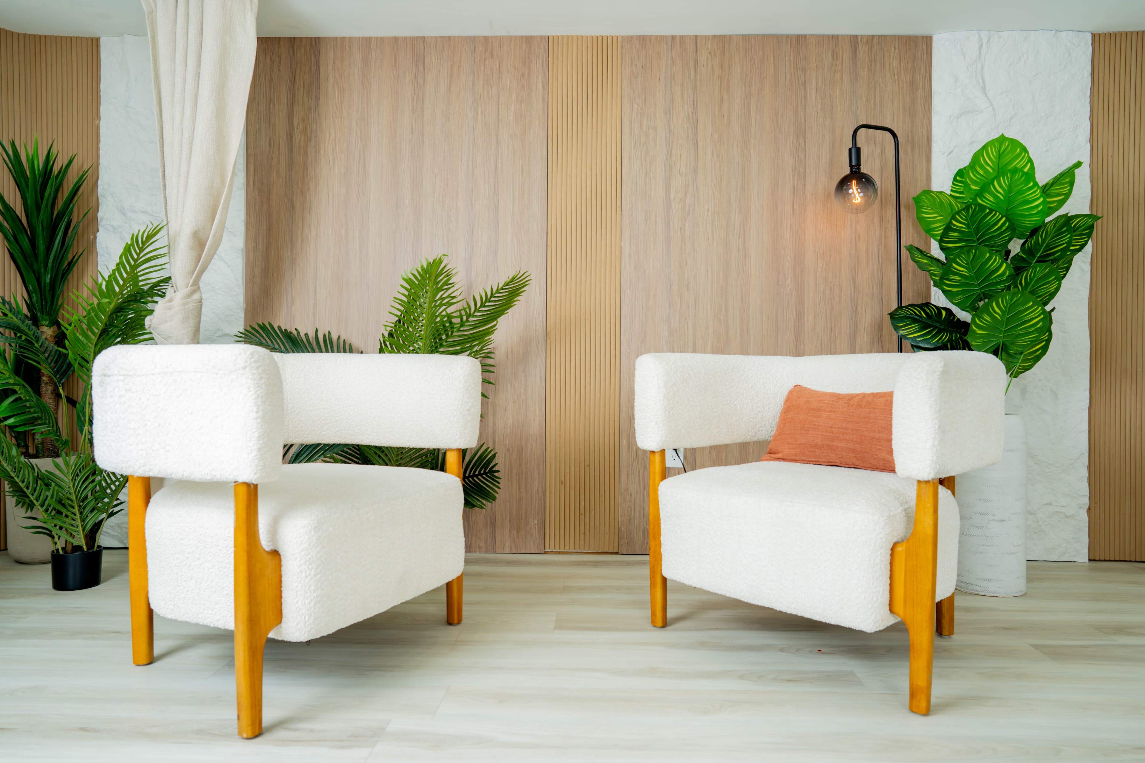 The image shows two white, upholstered chairs with wooden legs positioned in a bright, modern interior featuring plants and decorative wall panels.