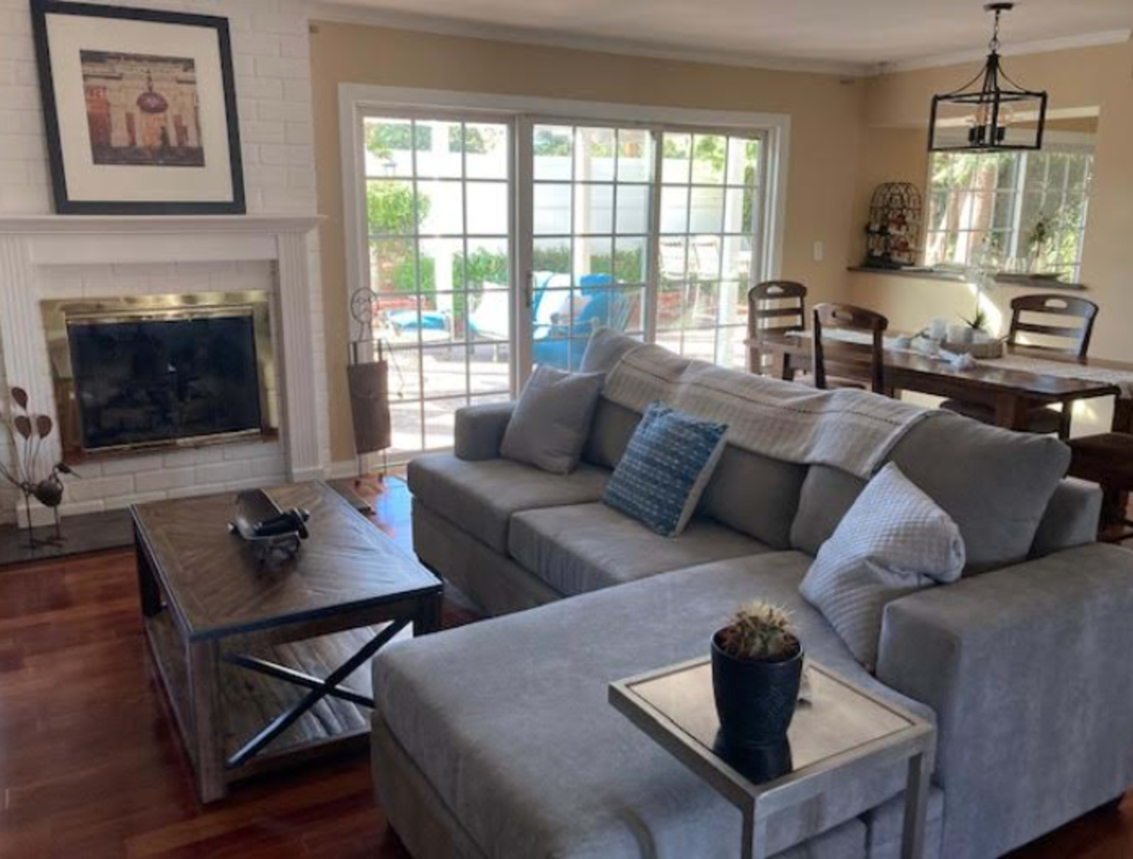 A living room with a gray sectional sofa, a wooden coffee table, and a fireplace, with large windows that provide a view of an outdoor patio.