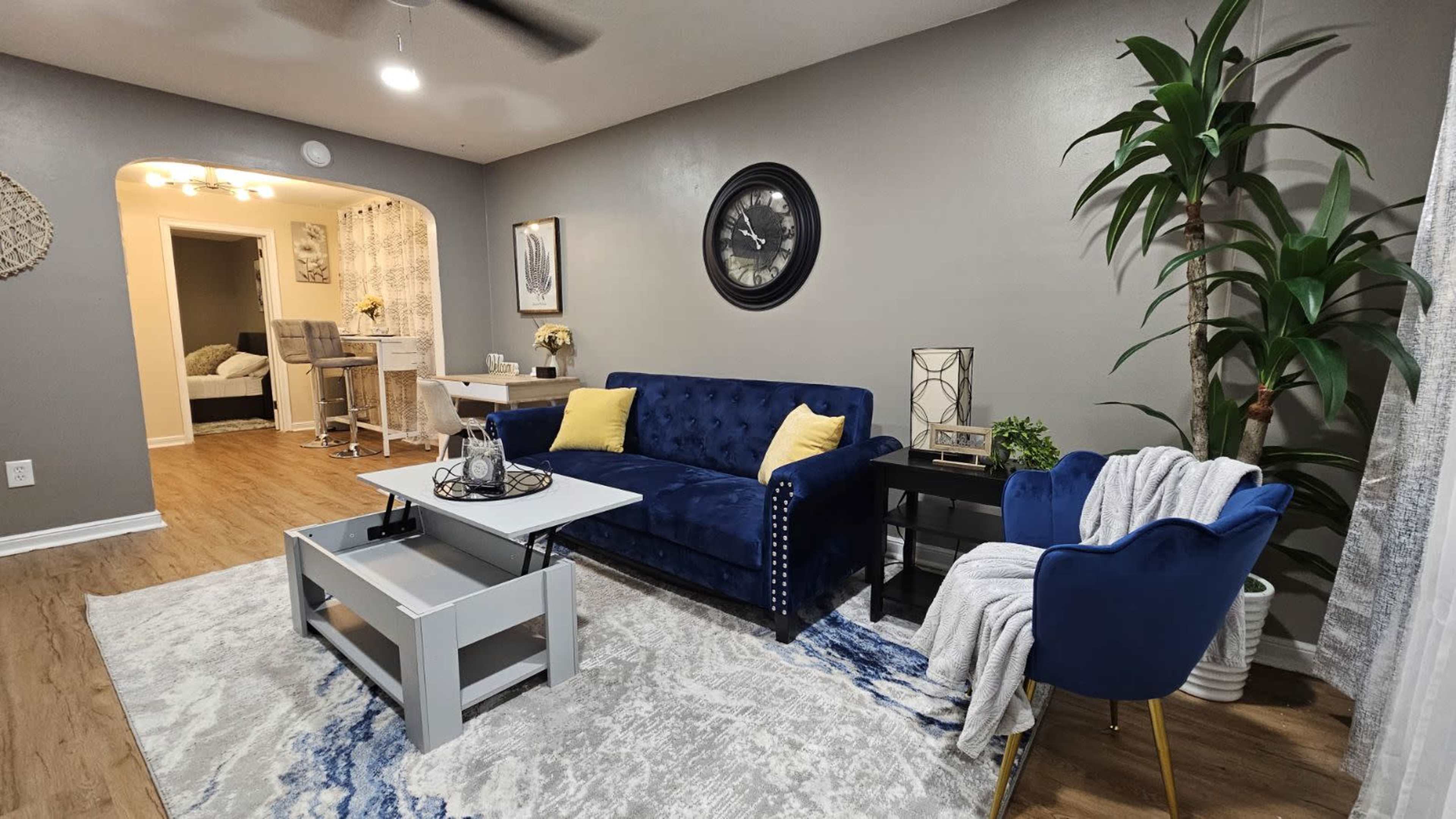 A modern living room featuring a blue velvet sofa, a light gray coffee table, a decorative plant, and a prominent wall clock.