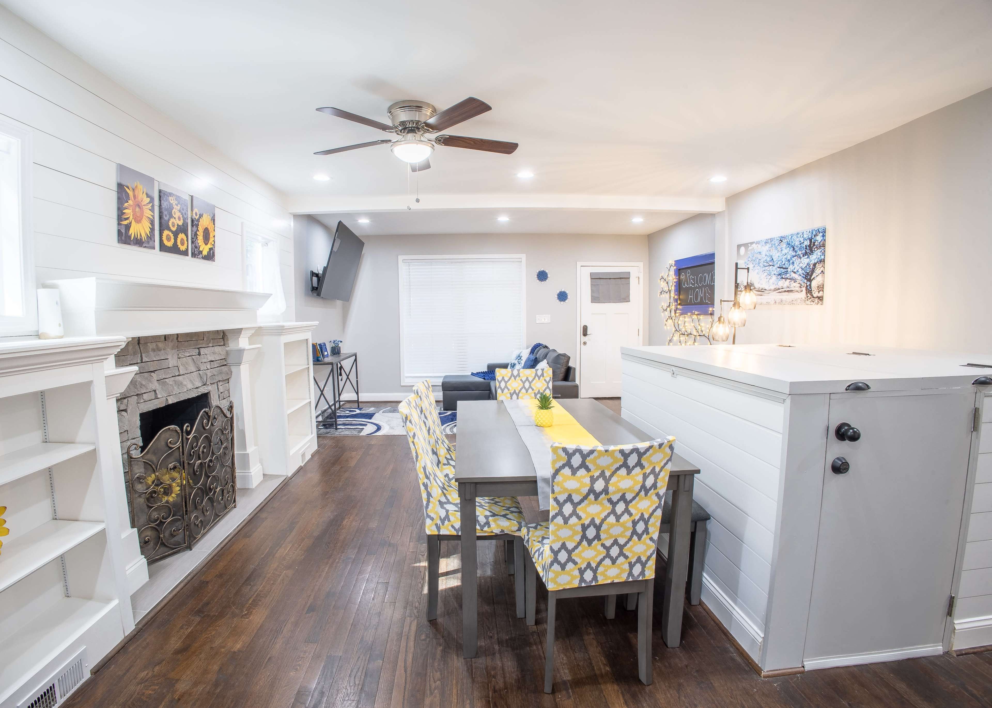 A living room and dining area featuring a stone fireplace, a table with yellow and blue patterned chairs, and a television mounted on the wall.