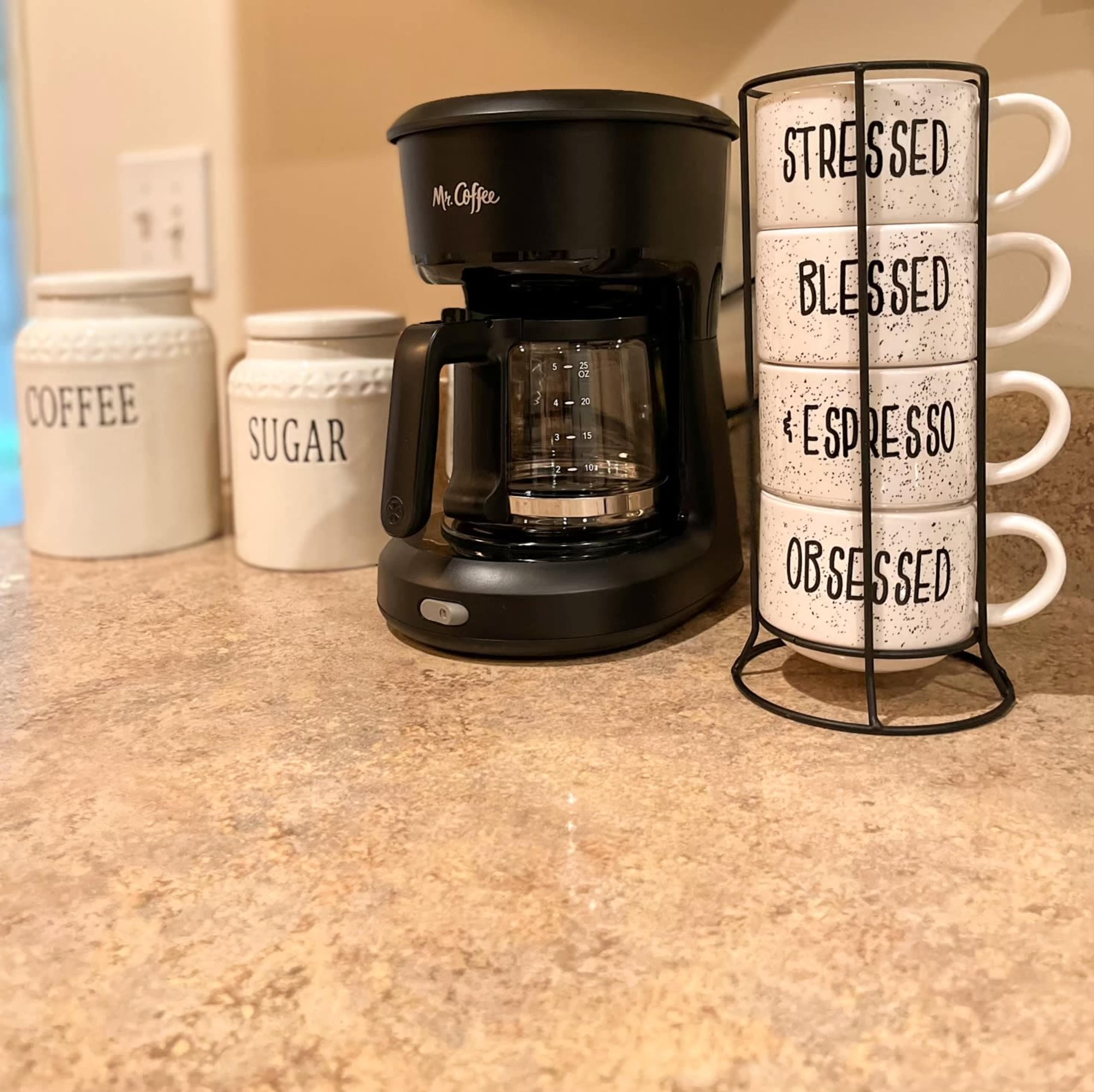 A coffee maker sits on a countertop next to decorative jars labeled "COFFEE" and "SUGAR," with a stacked holder of mugs displaying the words "STRESSED," "BLESSED," "EXPRESSO," and "OBSESSED."