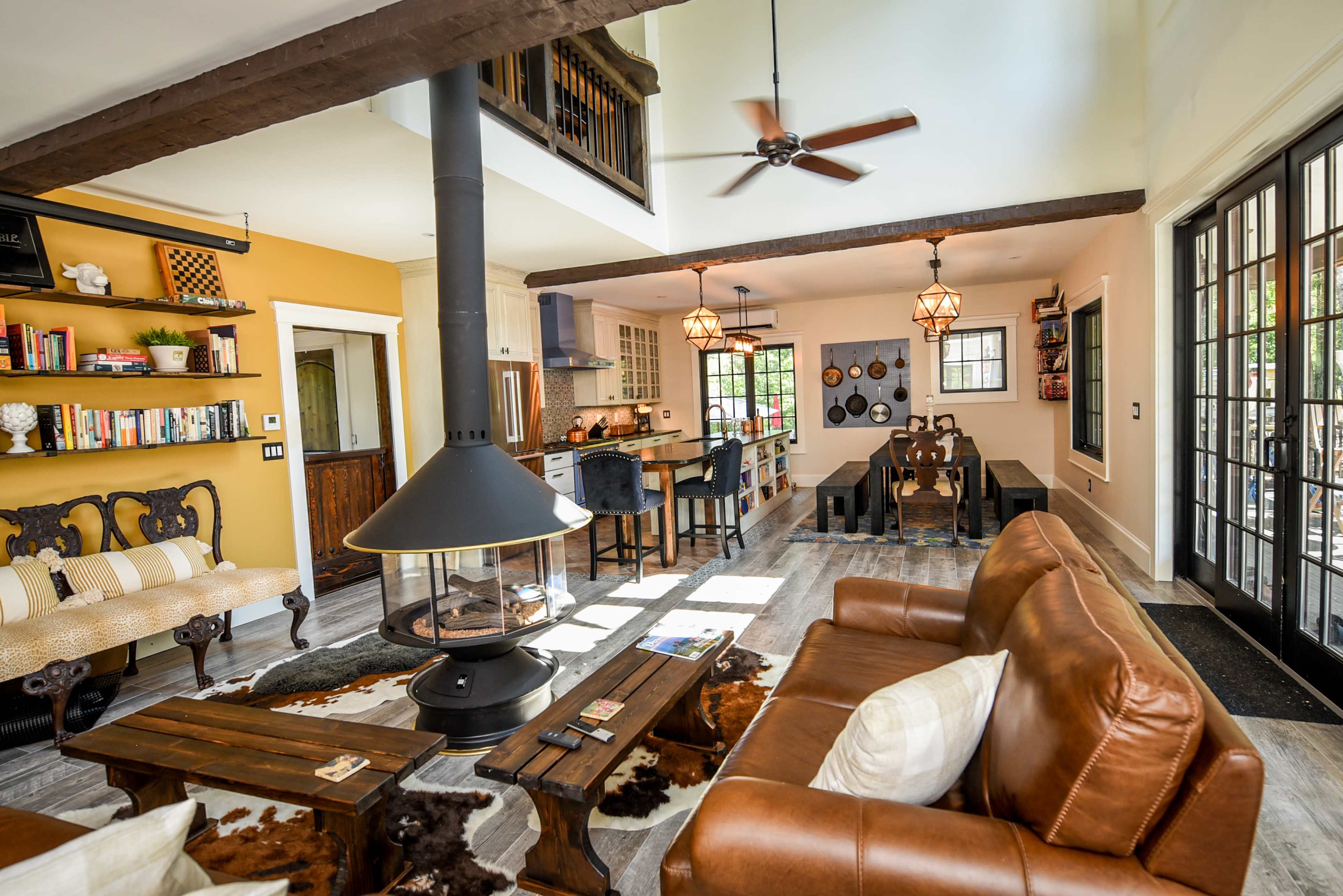A living area features a brown leather sofa, a central fireplace, and an open layout connecting a dining space and kitchen with wooden beams and warm-colored walls.