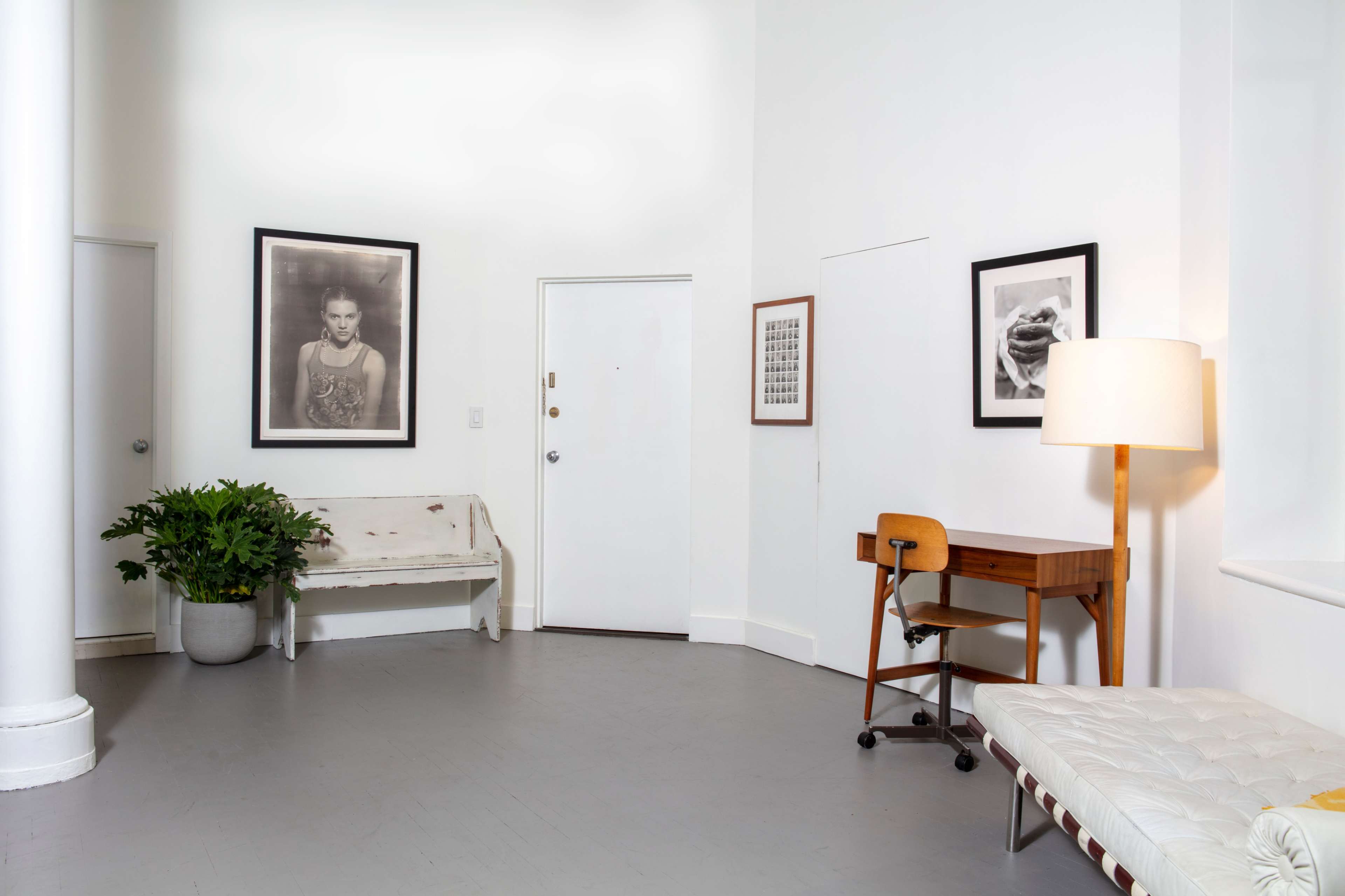 A minimalist room containing a bench, a wooden desk with a chair, framed photographs on the walls, and a floor lamp beside a potted plant.