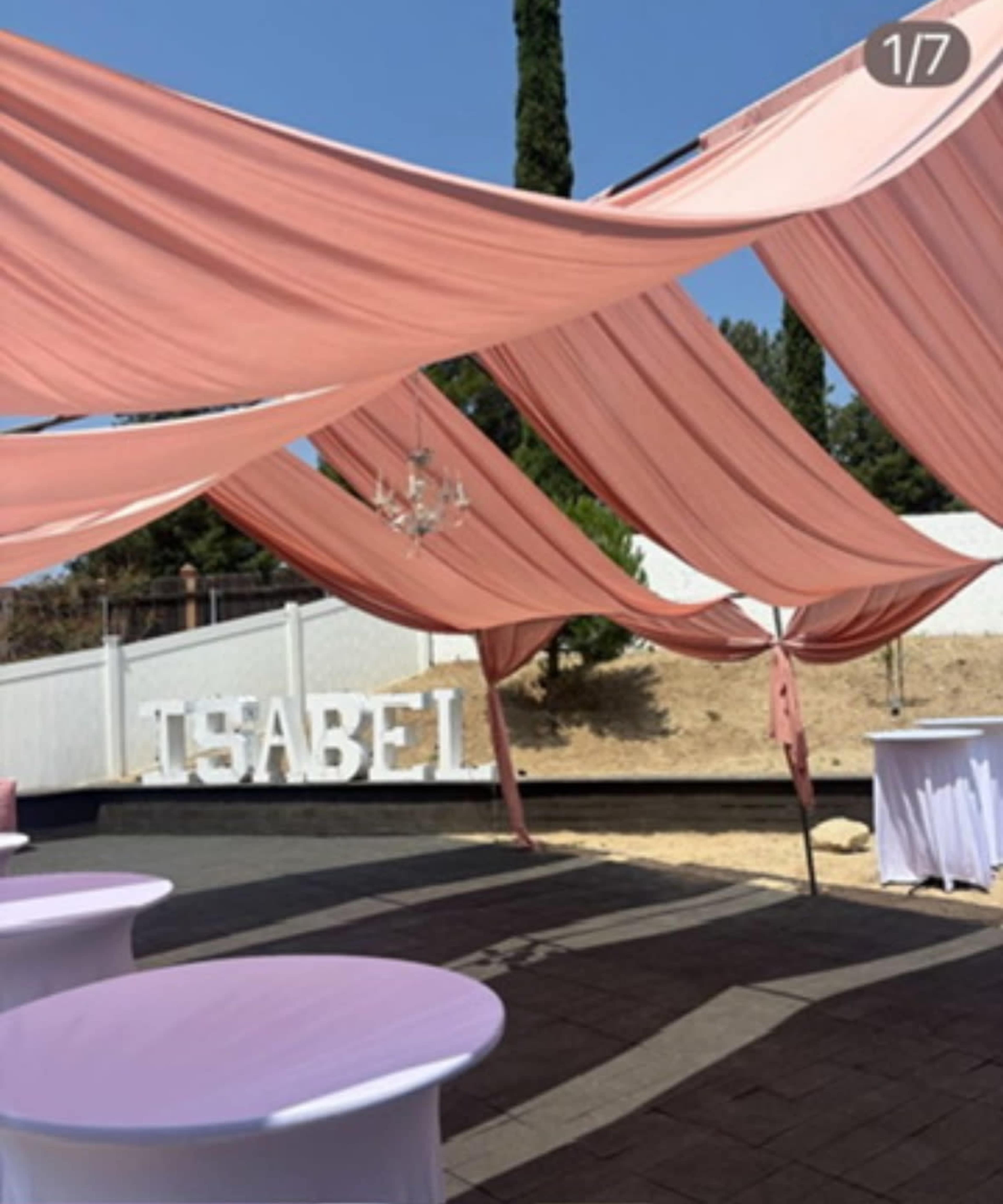 The image shows an outdoor event space decorated with draped pink fabric, white tables, and large letters spelling "ISABEL" against a backdrop of trees and a clear sky.