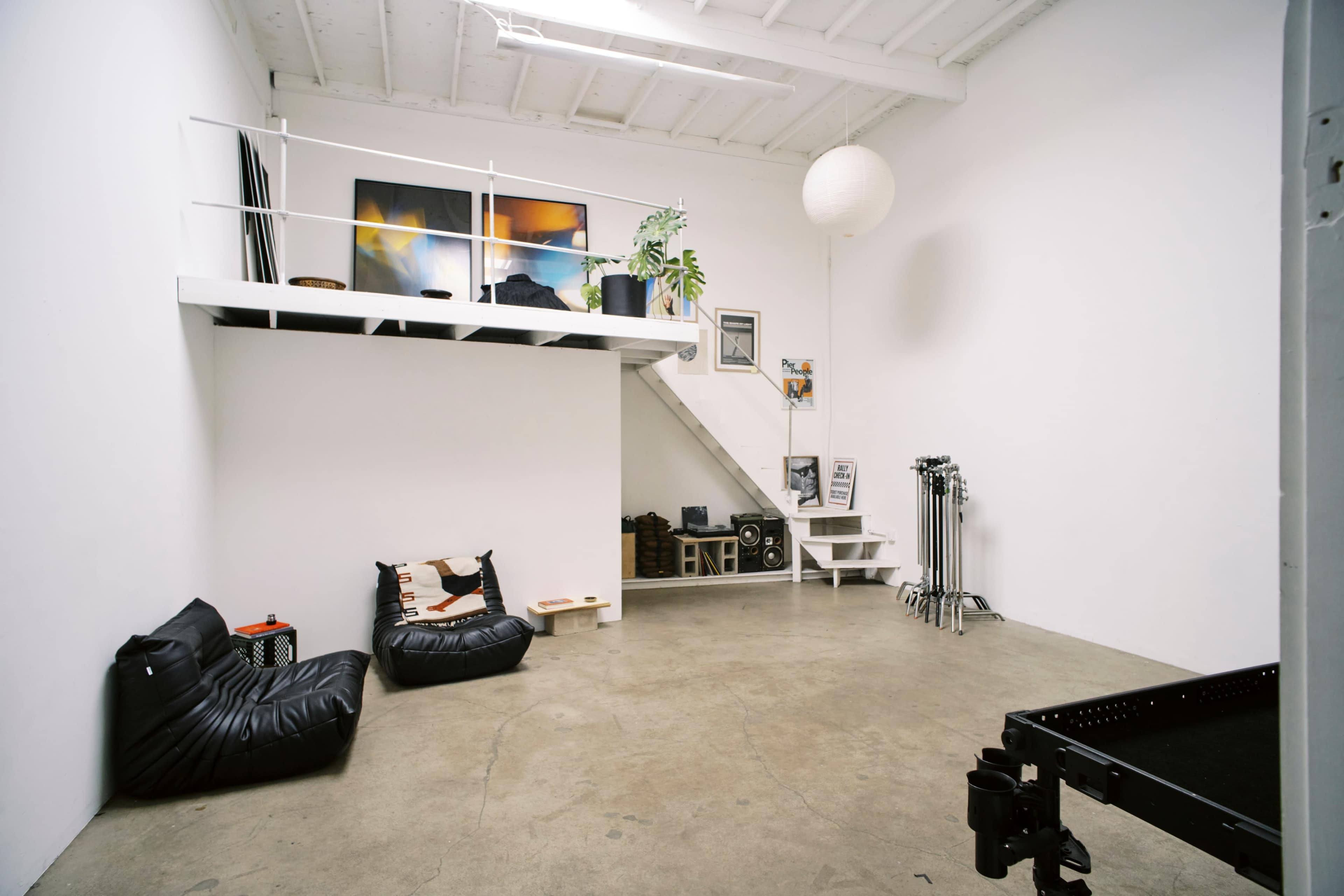 The image shows a minimalist interior space with a loft area, bean bag seating, and various art and equipment arranged around the room.